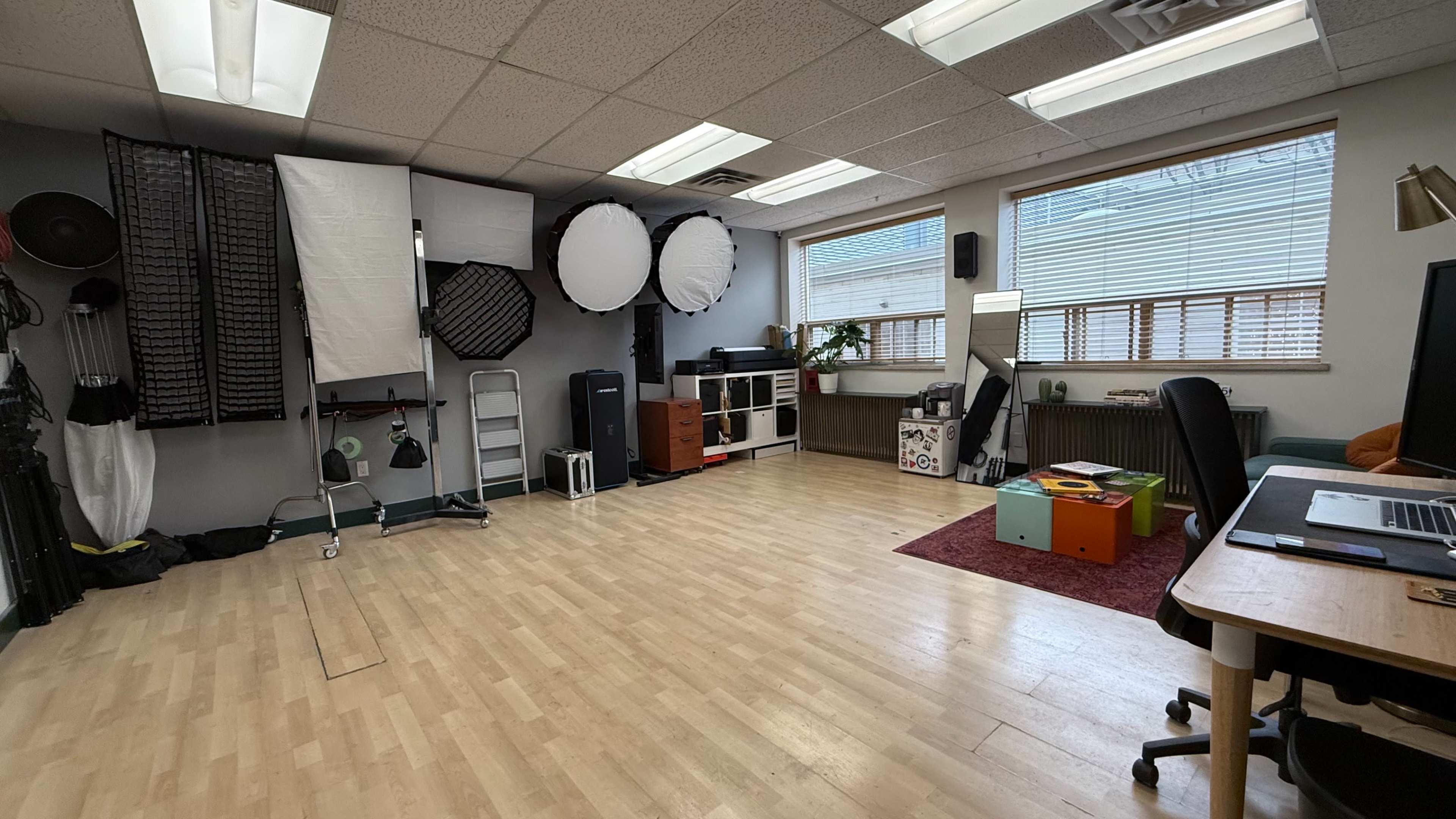 The image shows a spacious photography studio with various lighting equipment and softboxes mounted on the walls, wooden flooring, and a desk setup with a computer.