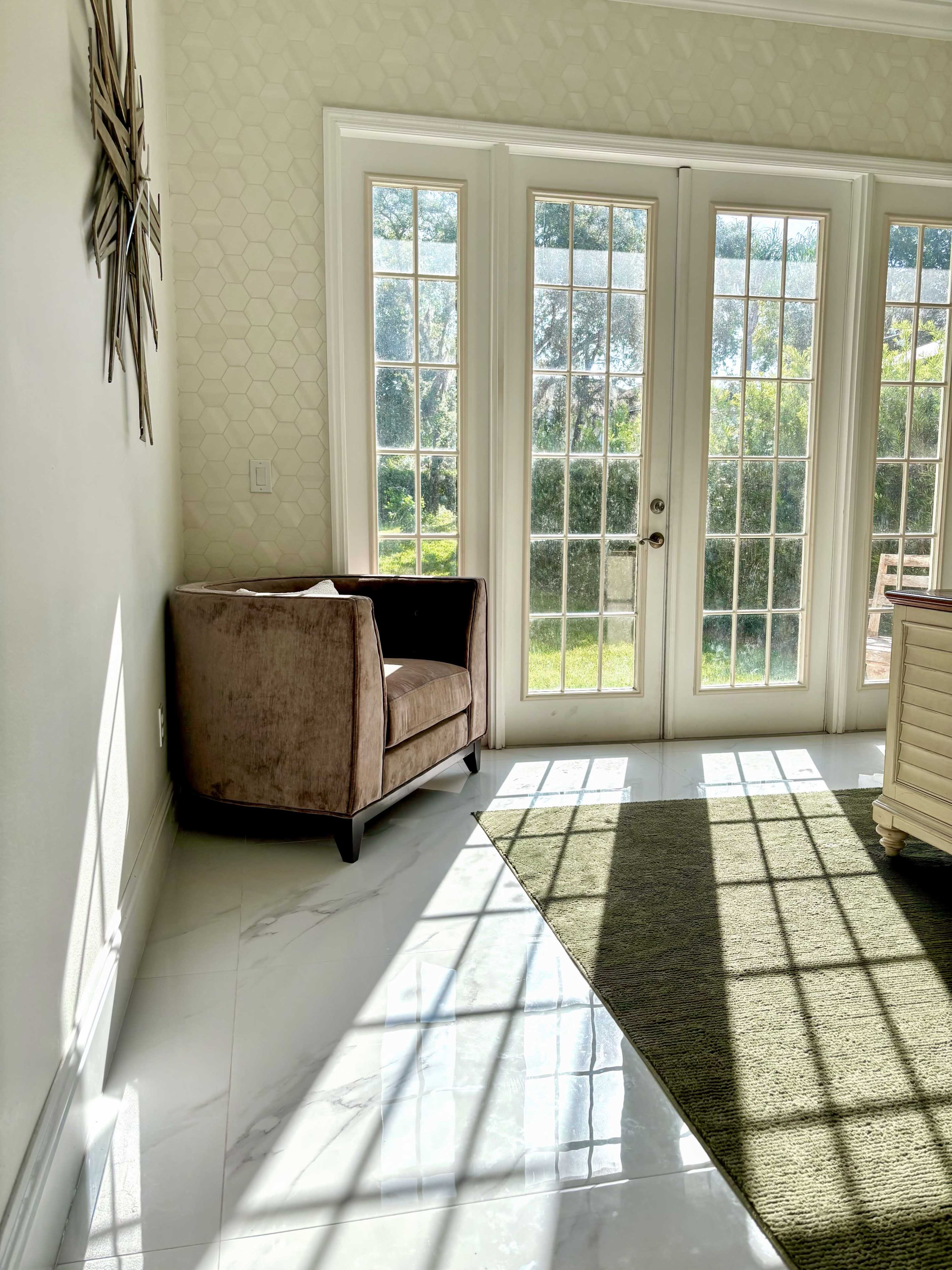A brown armchair is positioned near large, glass doors that lead to a garden, with sunlight casting geometric shadows on the floor.