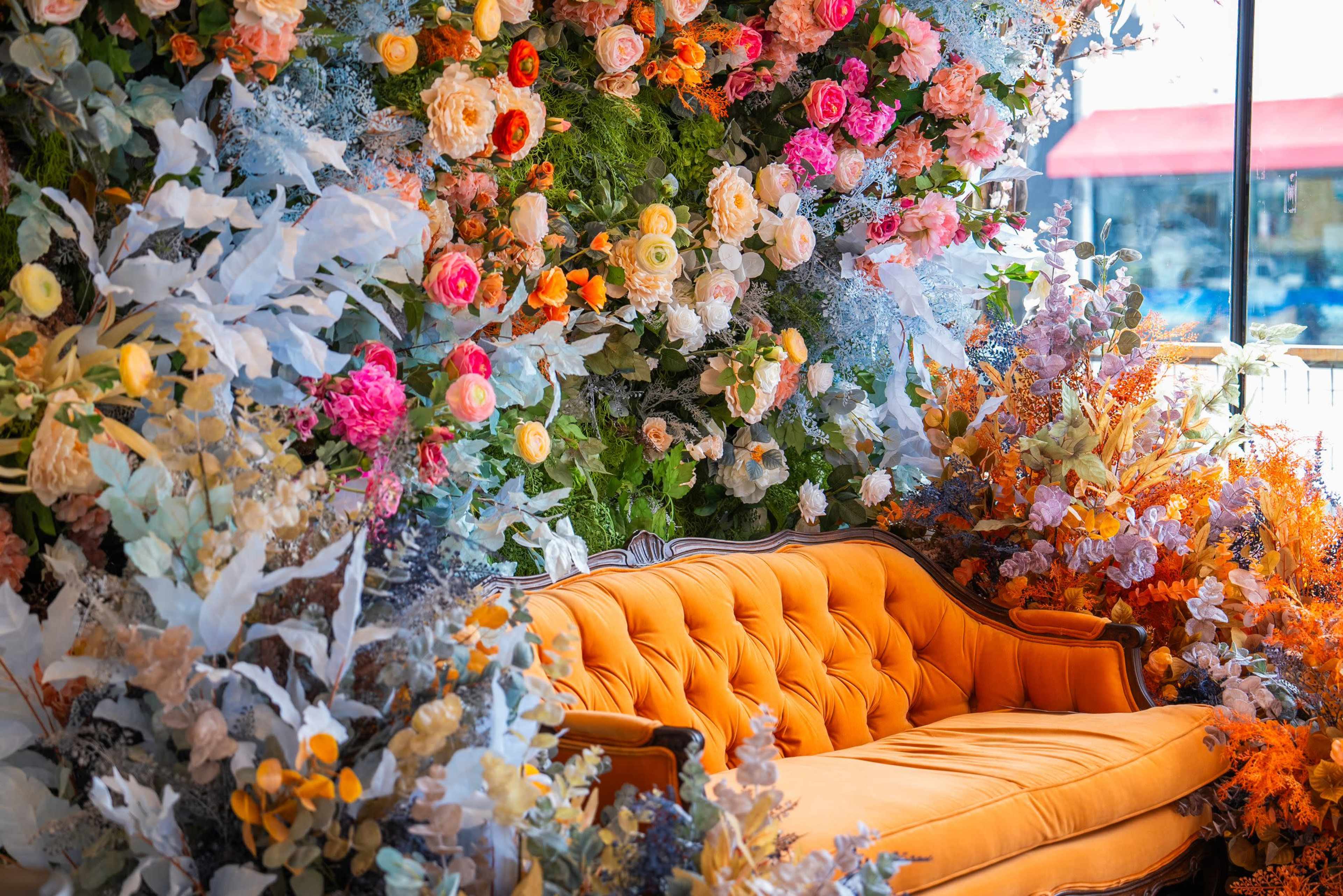 A vintage orange sofa is positioned in front of a colorful floral wall display featuring various artificial flowers and greenery.