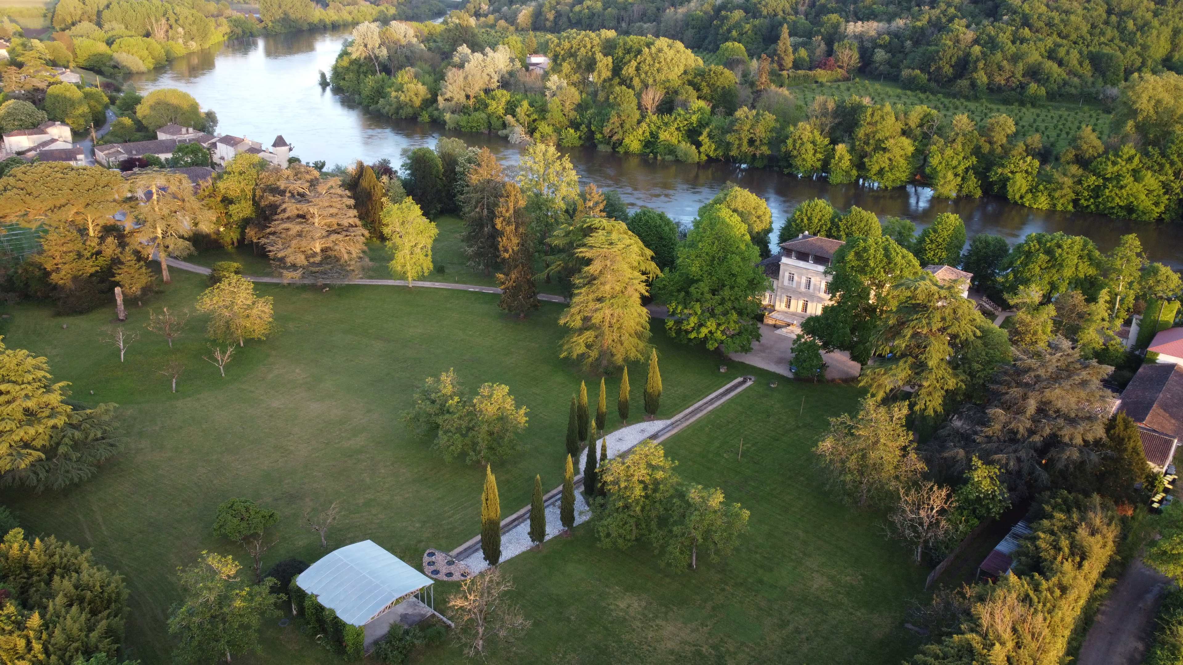 The image shows a scenic landscape featuring a river surrounded by lush greenery, with a manicured lawn and a pathway leading to a house.