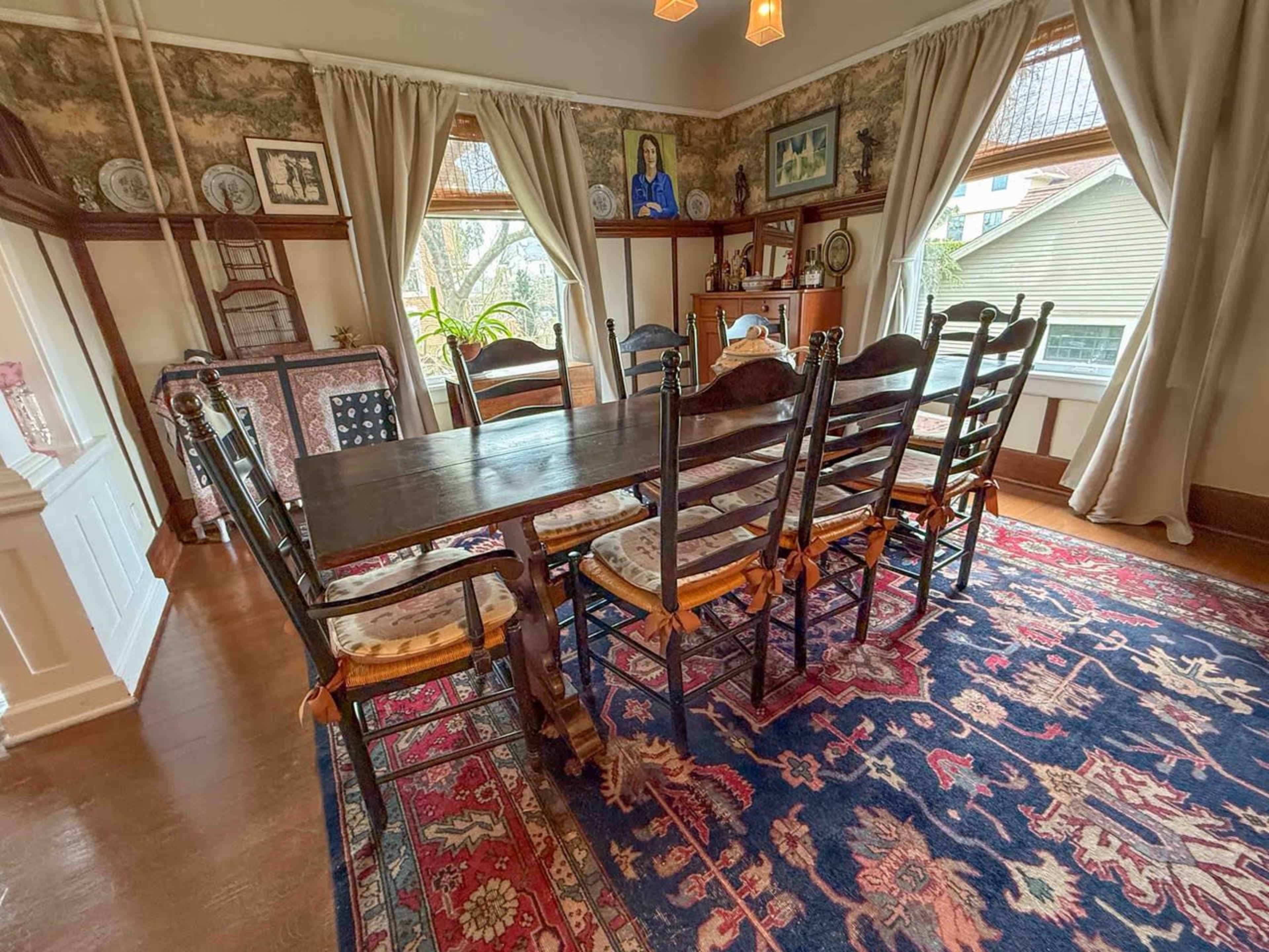 A long wooden dining table surrounded by eight chairs is set in a warmly decorated dining room with large windows and a colorful rug.