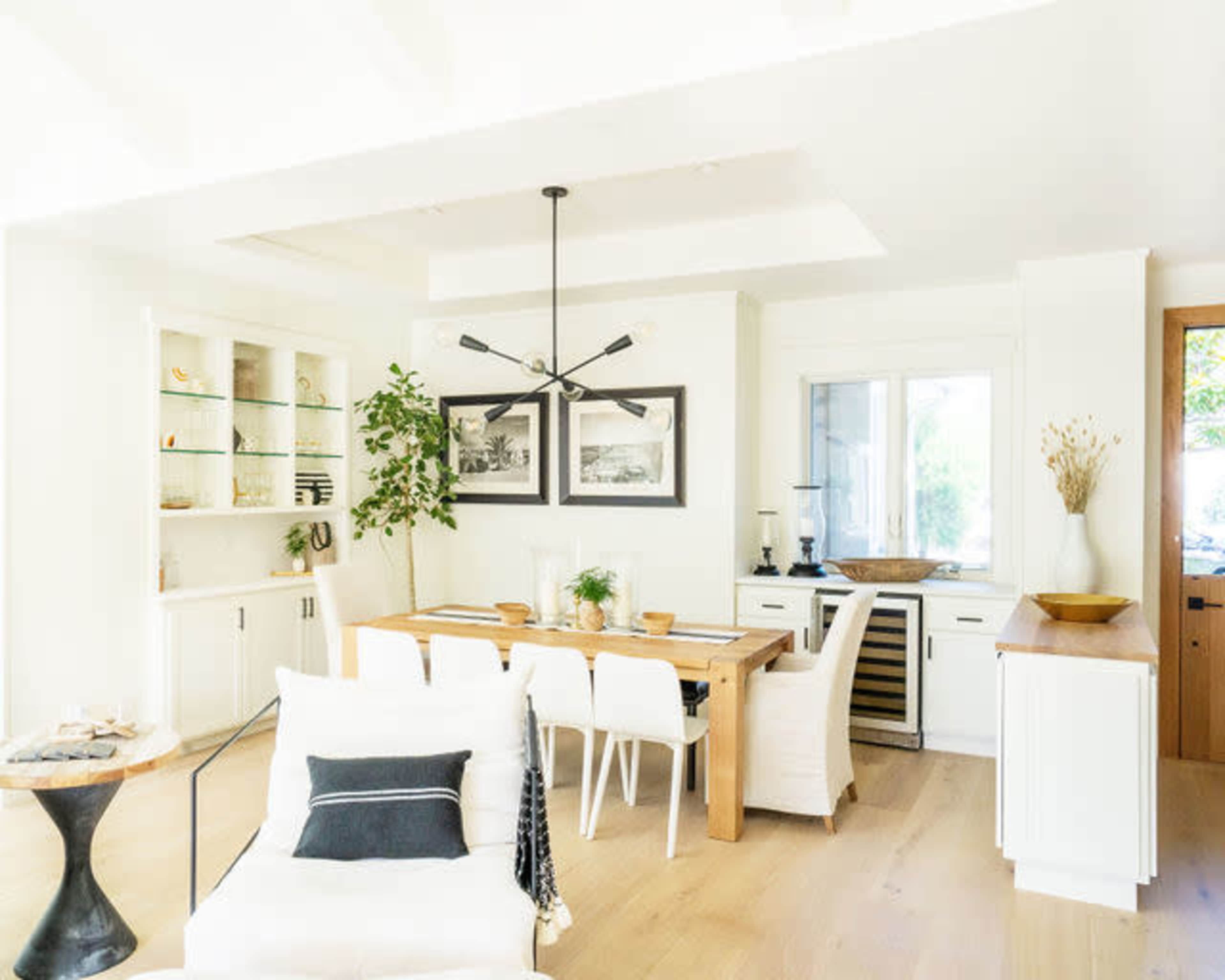 A bright dining area featuring a wooden table surrounded by white chairs, complemented by shelves and a window with natural decor elements.
