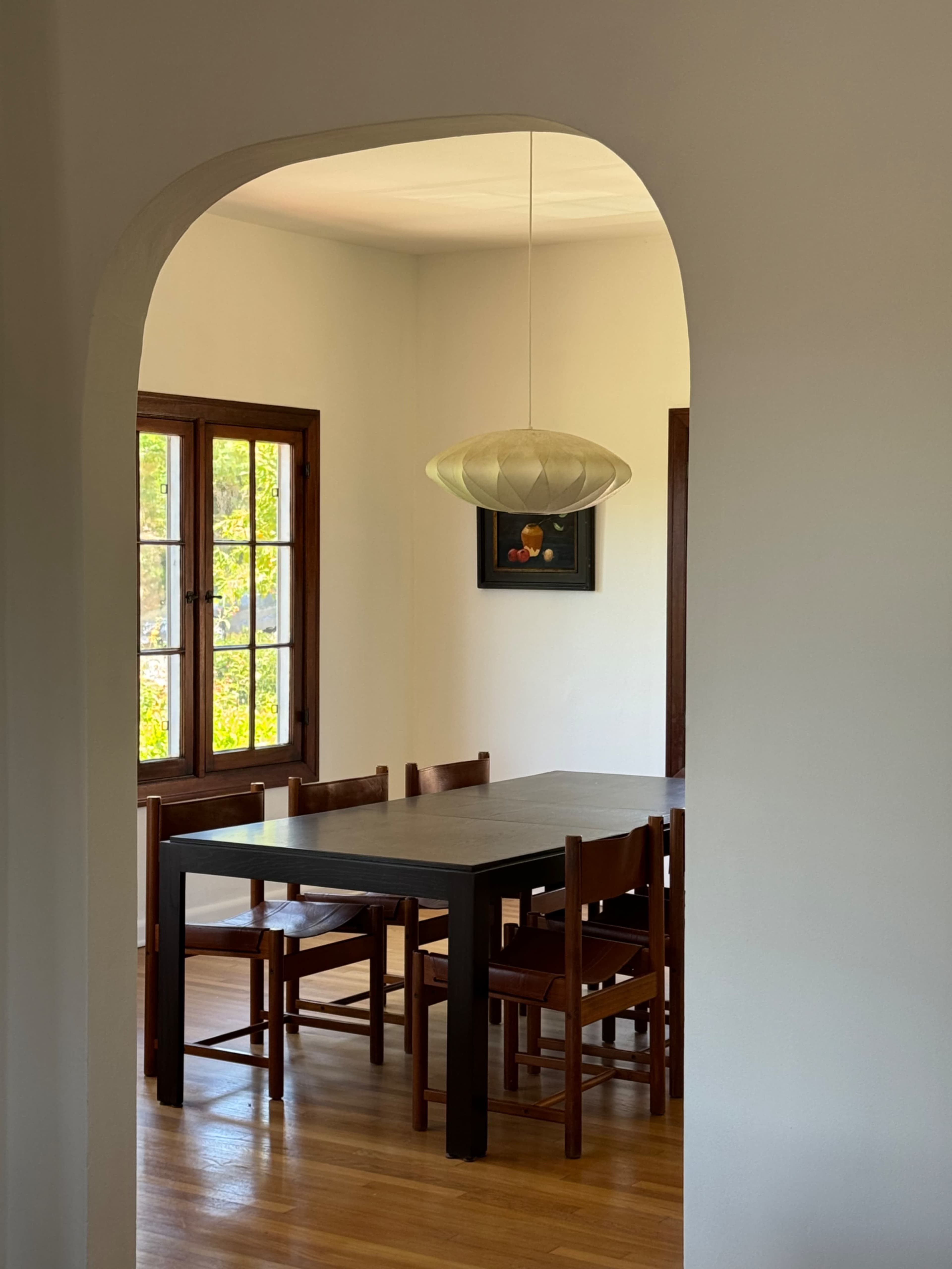 A dining room features a table surrounded by wooden chairs, illuminated by a pendant light, with large windows letting in natural light.