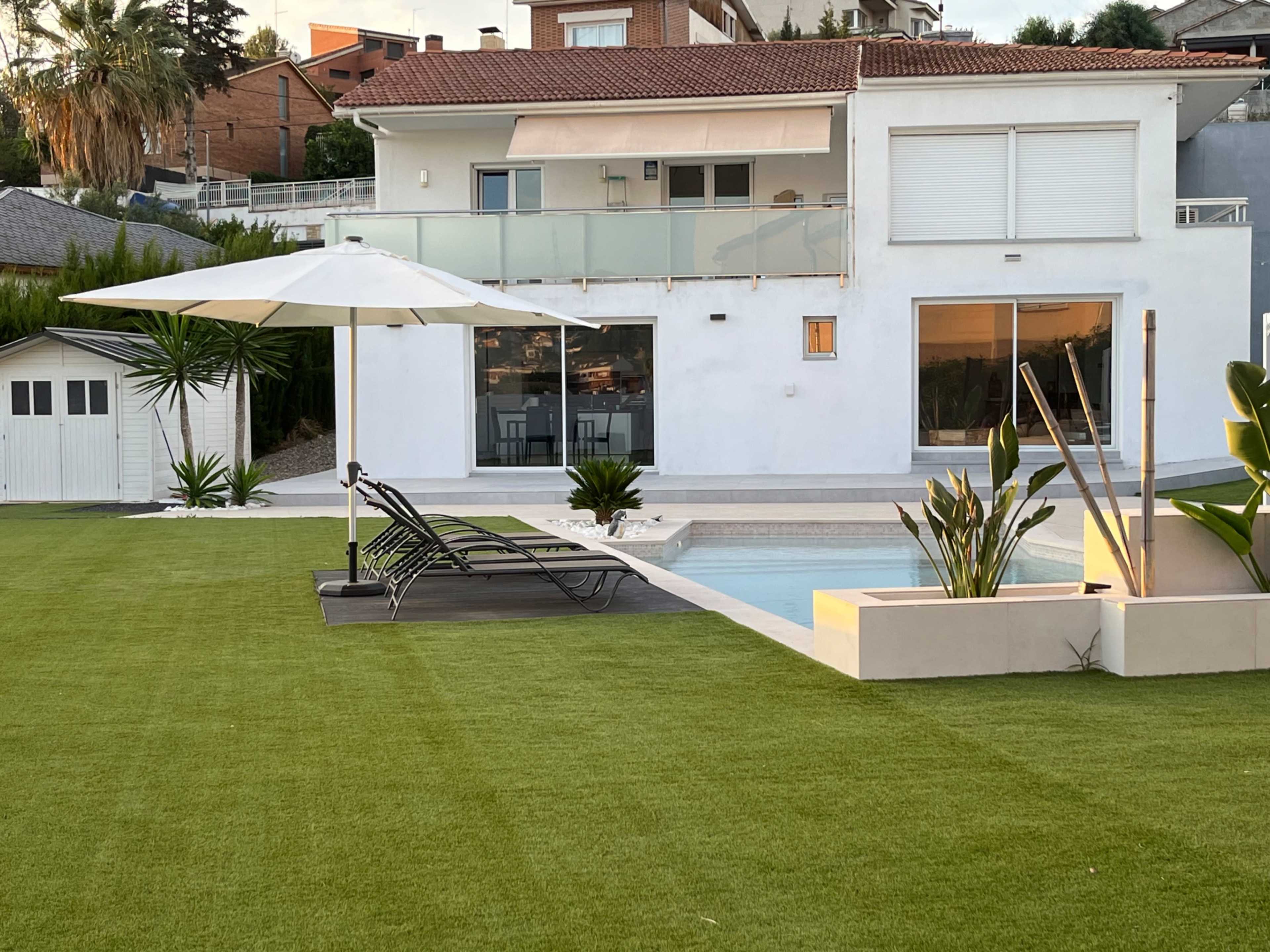 A modern white house with large windows, a pool, and a spacious lawn features a lounge chair under a parasol.