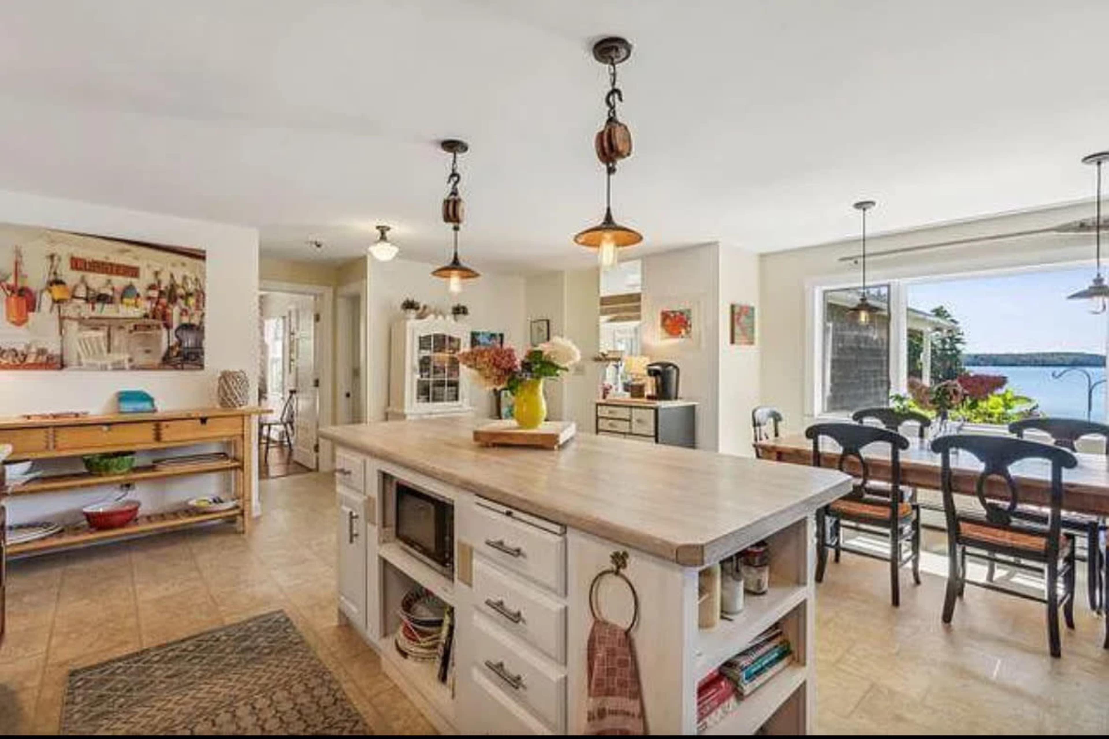 A bright kitchen features a central island with a countertop, wooden dining table by a window, and views of the water outside.