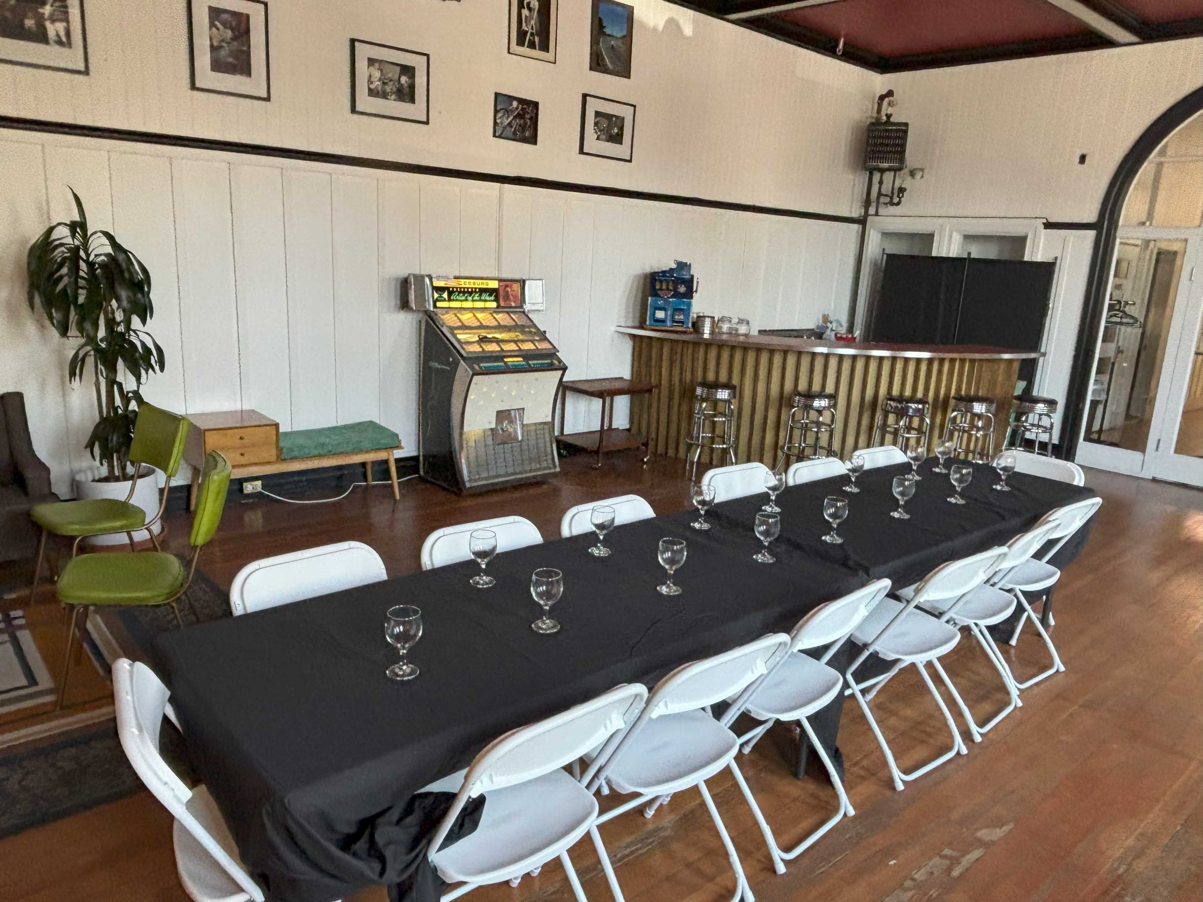A long black table with wine glasses is set up in a room featuring a jukebox, bar area, and framed photographs on the walls.