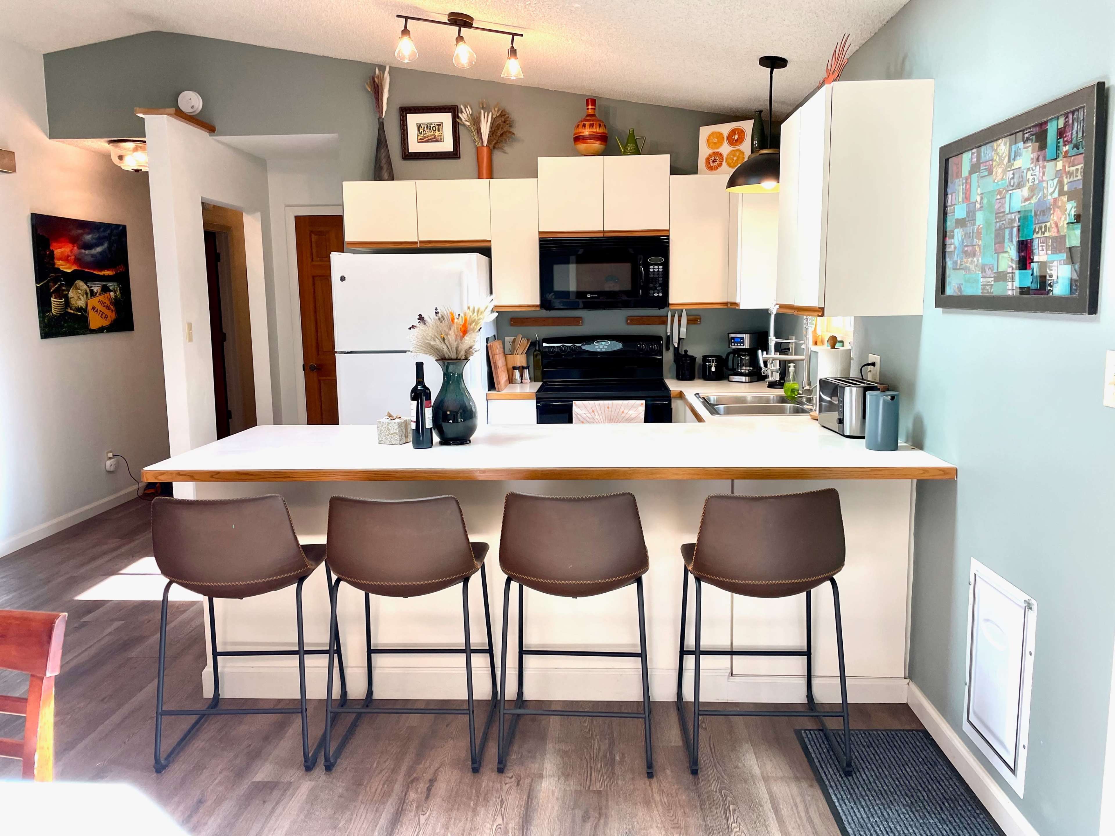 The image shows a modern kitchen with white cabinets, a black countertop, and four brown bar stools lined up at a kitchen island.