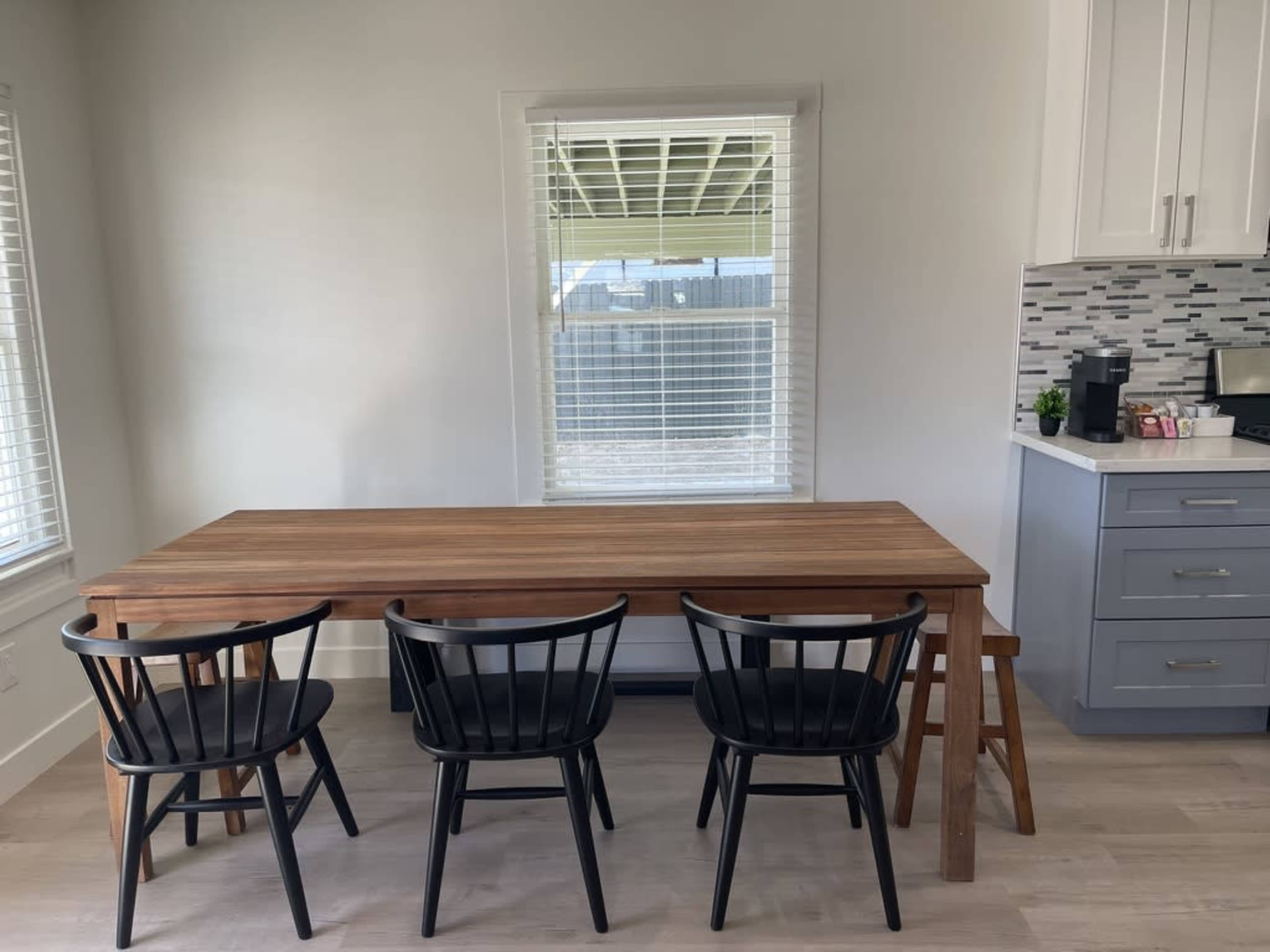 A wooden dining table with four black chairs is positioned near a window in a bright kitchen.
