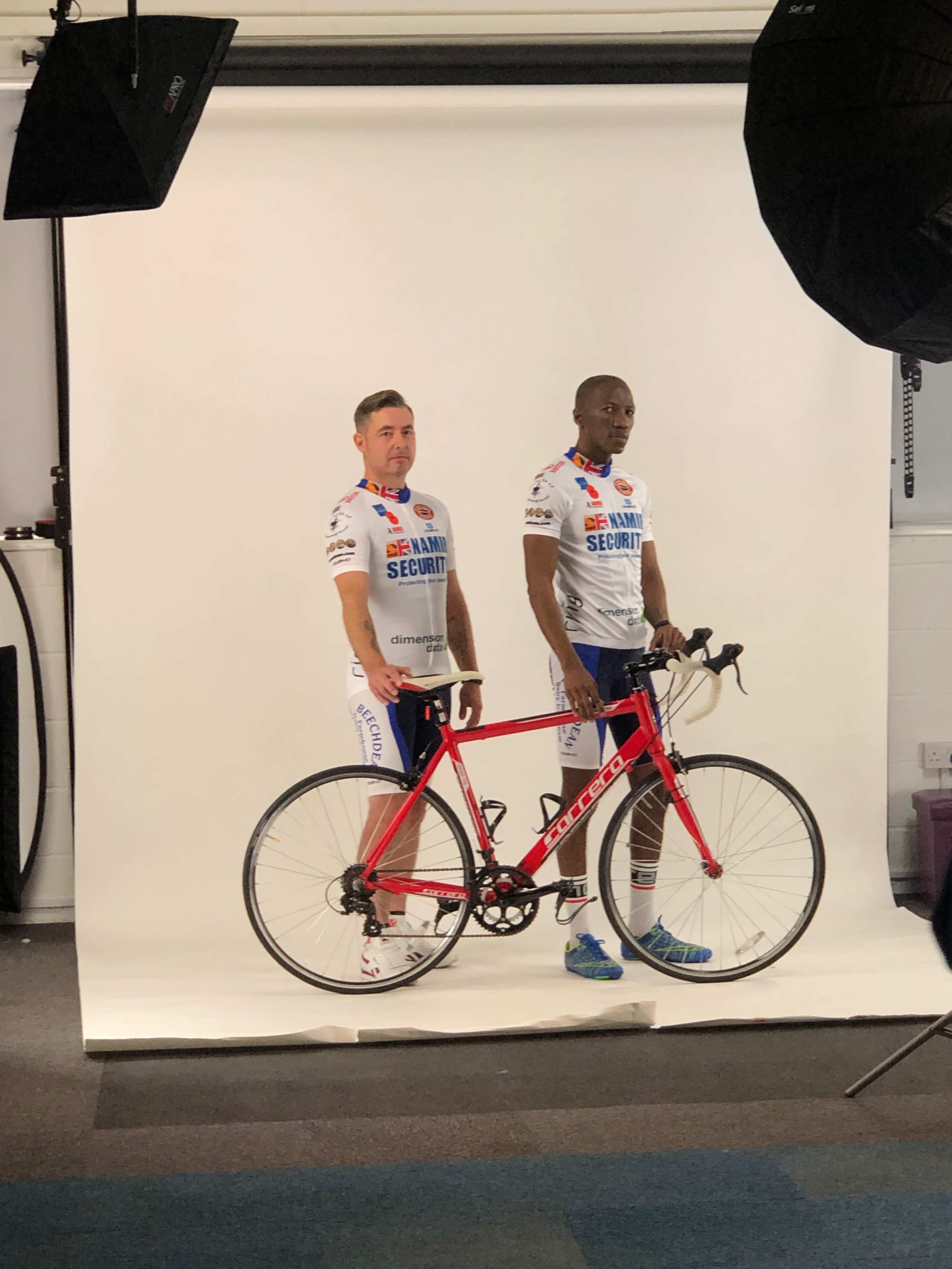 Two men in cycling jerseys stand beside a red bicycle in a studio setting with a plain backdrop.