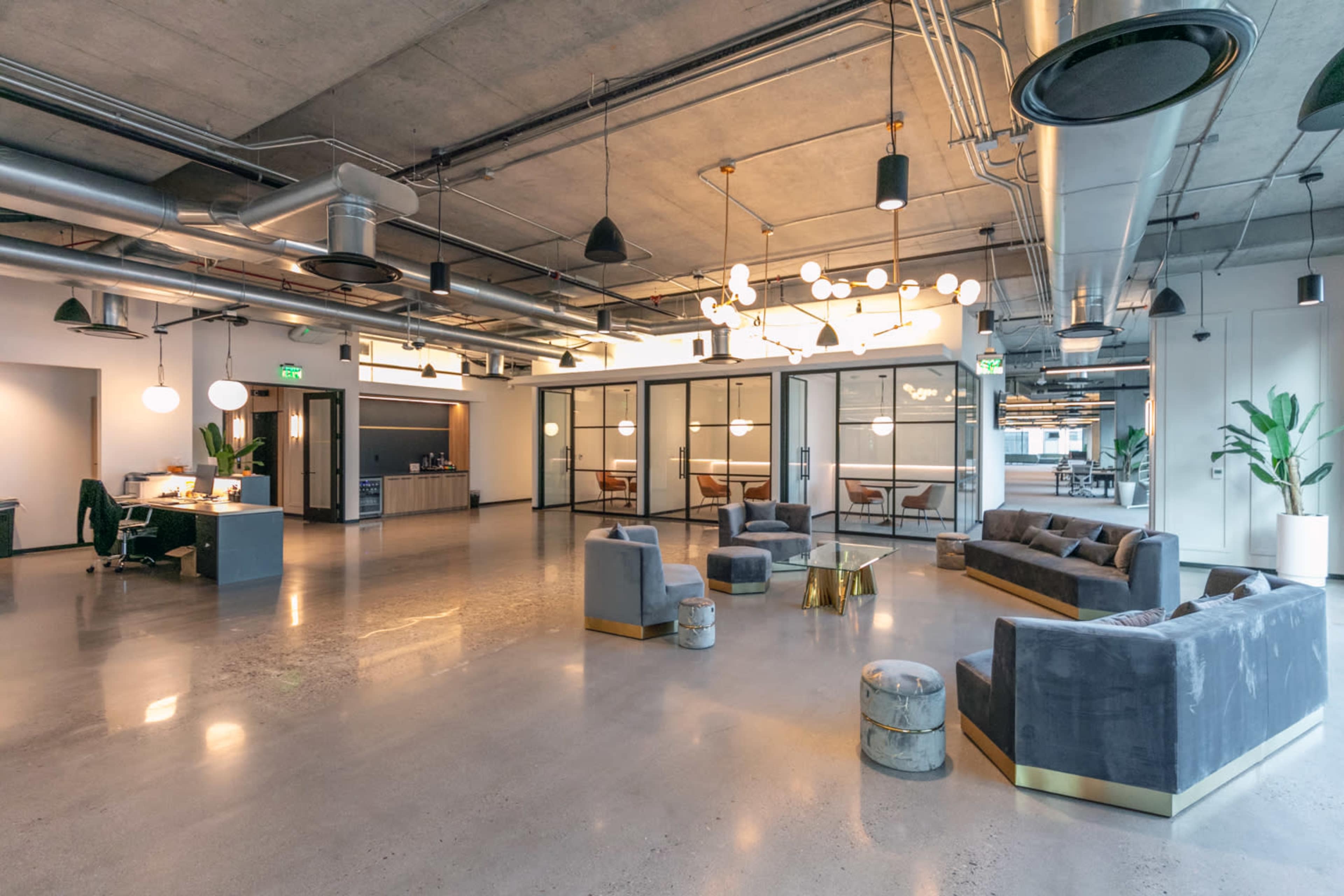 A modern office space features a seating area with gray sofas and decorative lighting, alongside glass-walled conference rooms and various potted plants.