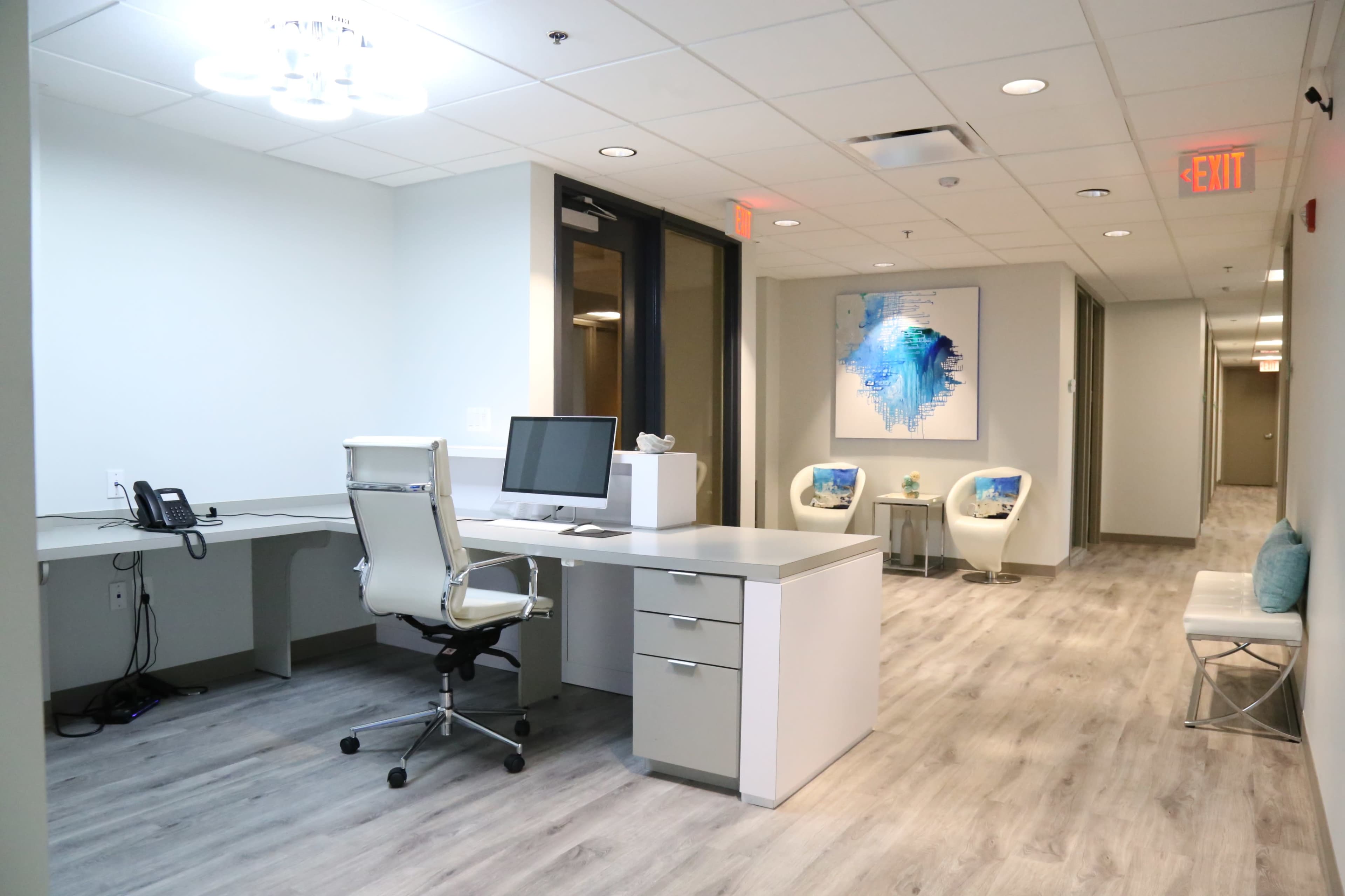 The image shows a modern office space featuring a desk with a computer, a chair, and two decorative chairs in a well-lit corridor.