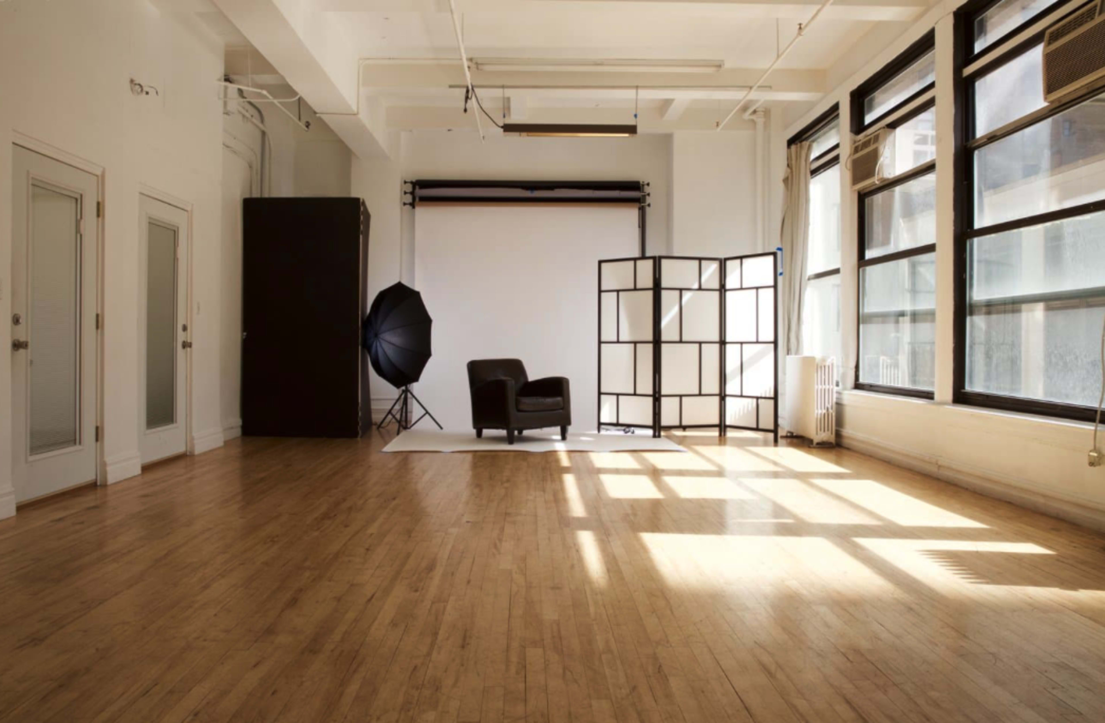 The image shows a spacious photography studio with wooden floors, large windows, a backdrop setup, and a black chair beside a folding screen.