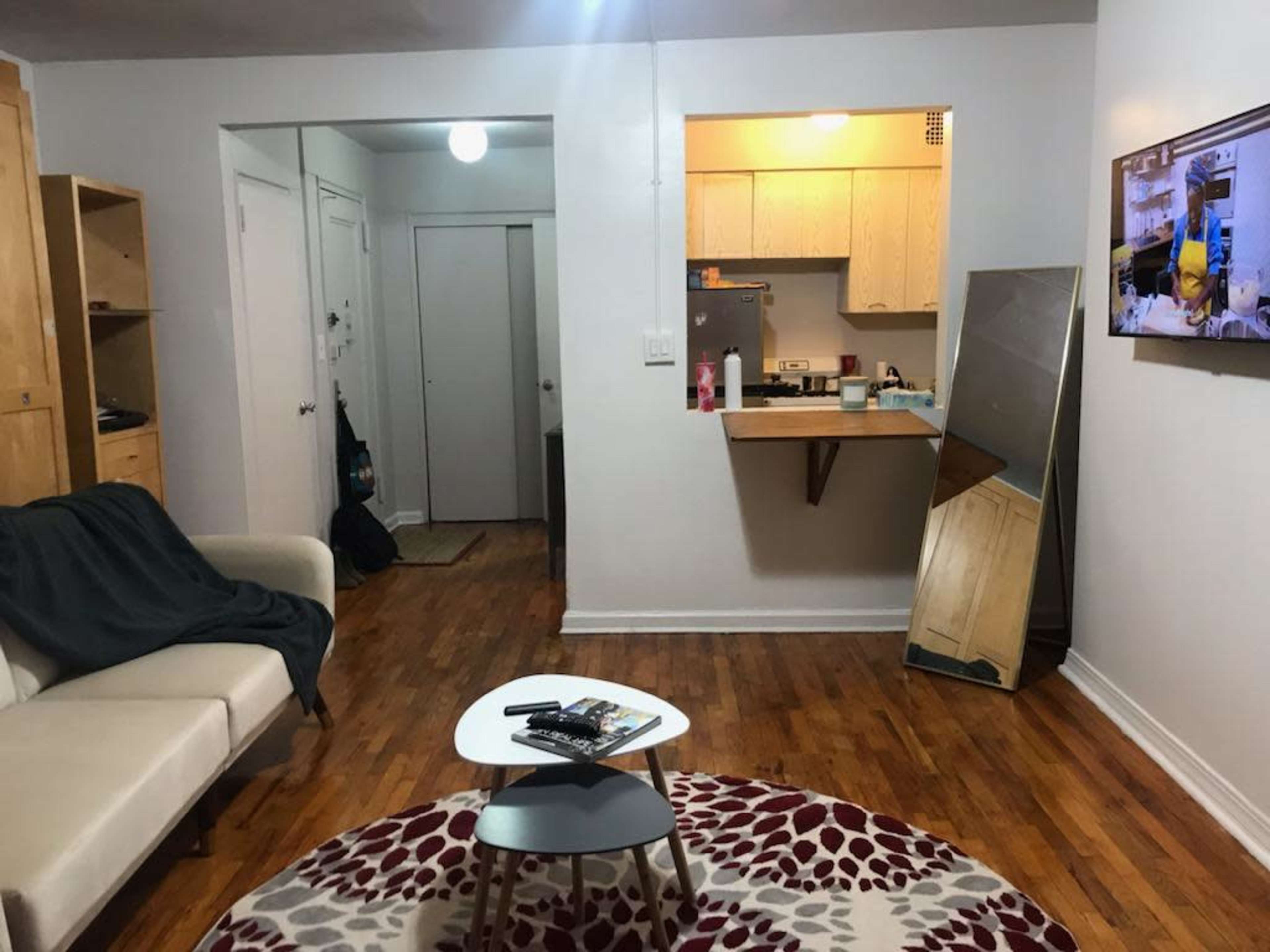 The image shows a small living area with a couch, a coffee table, a kitchen visible in the background, and a television mounted on the wall.