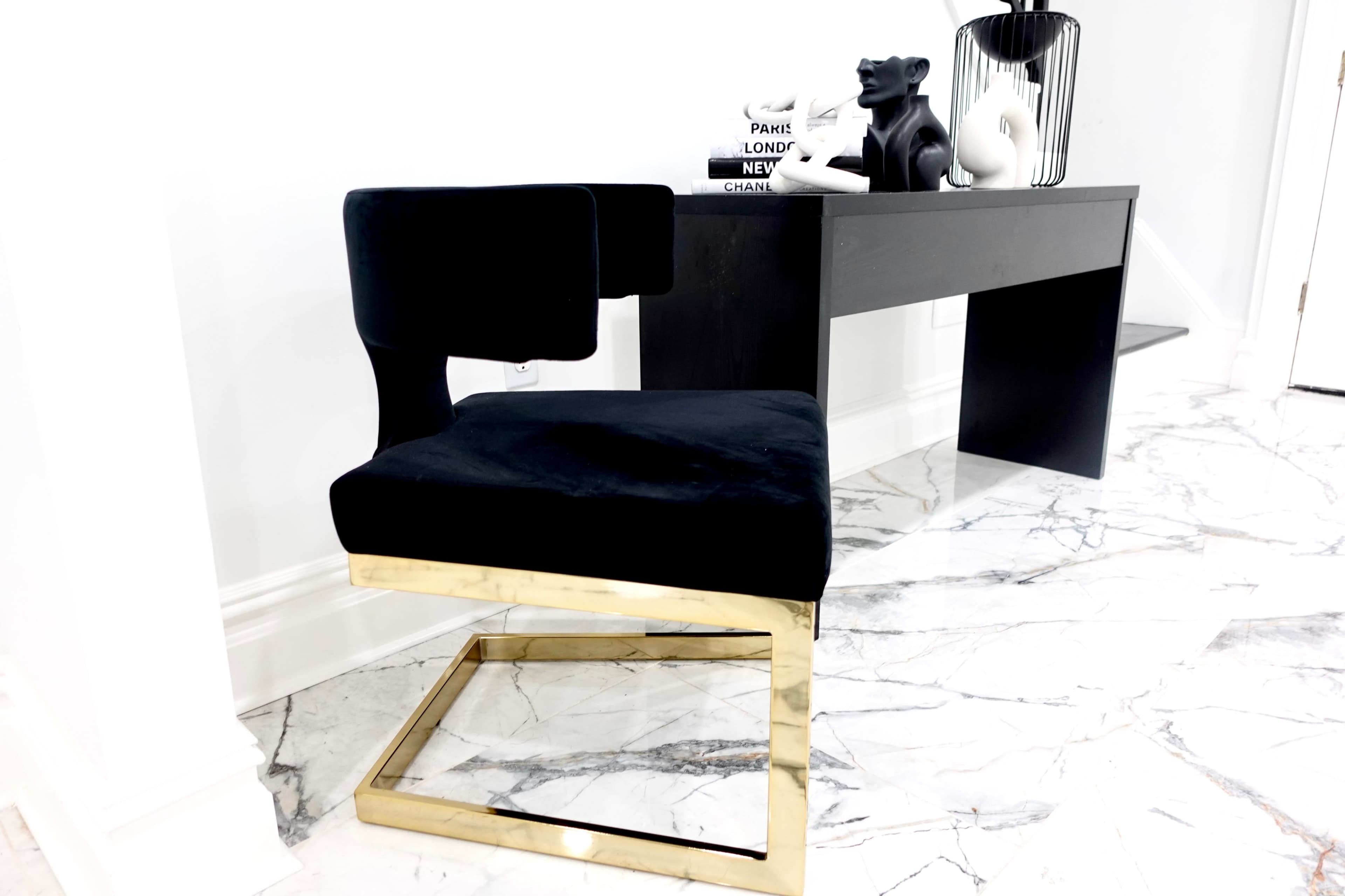 A black upholstered chair with a gold metal base is positioned next to a sleek black desk against a white wall, with decorative items on the desk and a marble floor beneath.