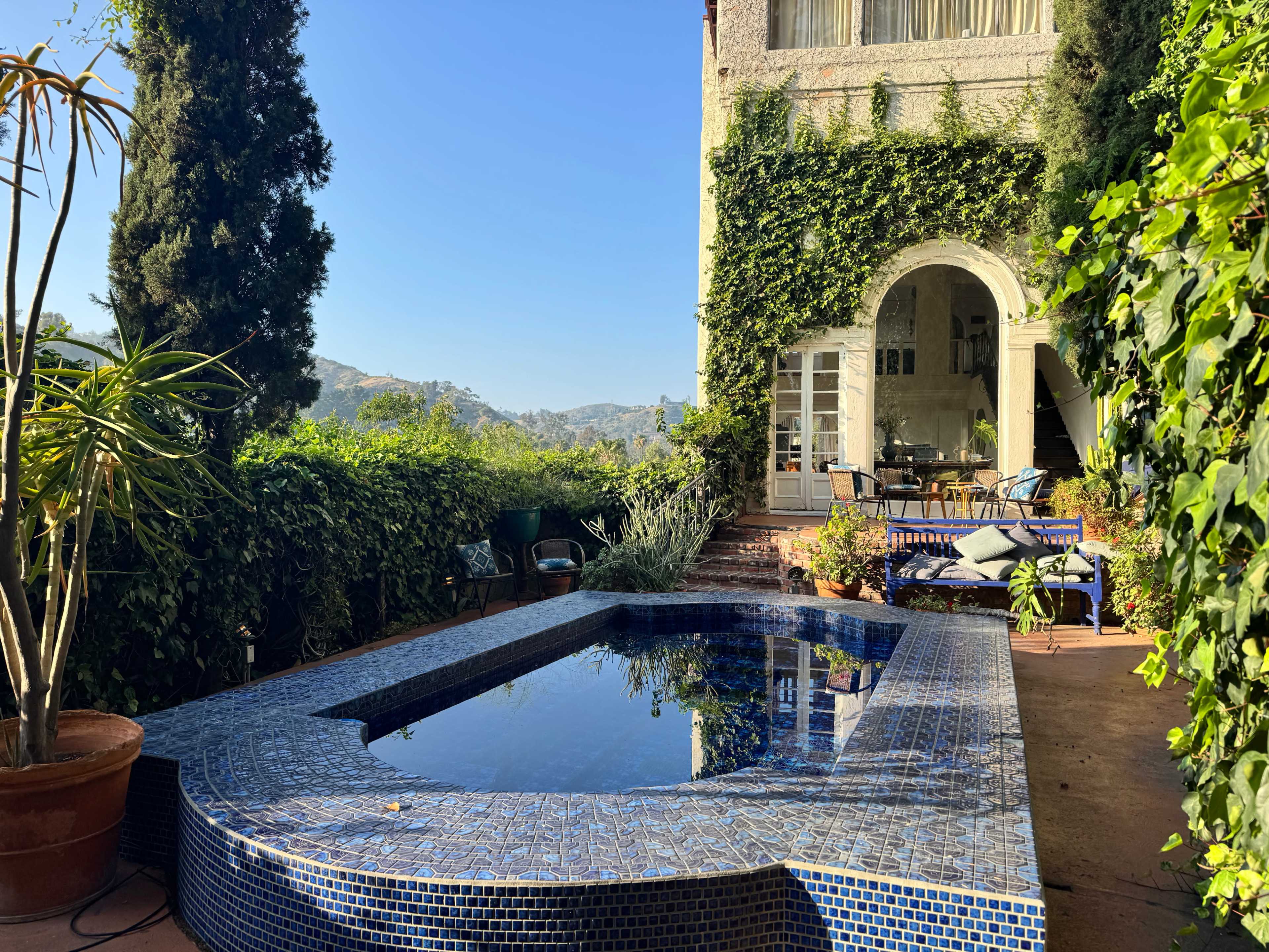 A serene outdoor area features a tiled pool bordered by lush greenery and a view of distant hills.