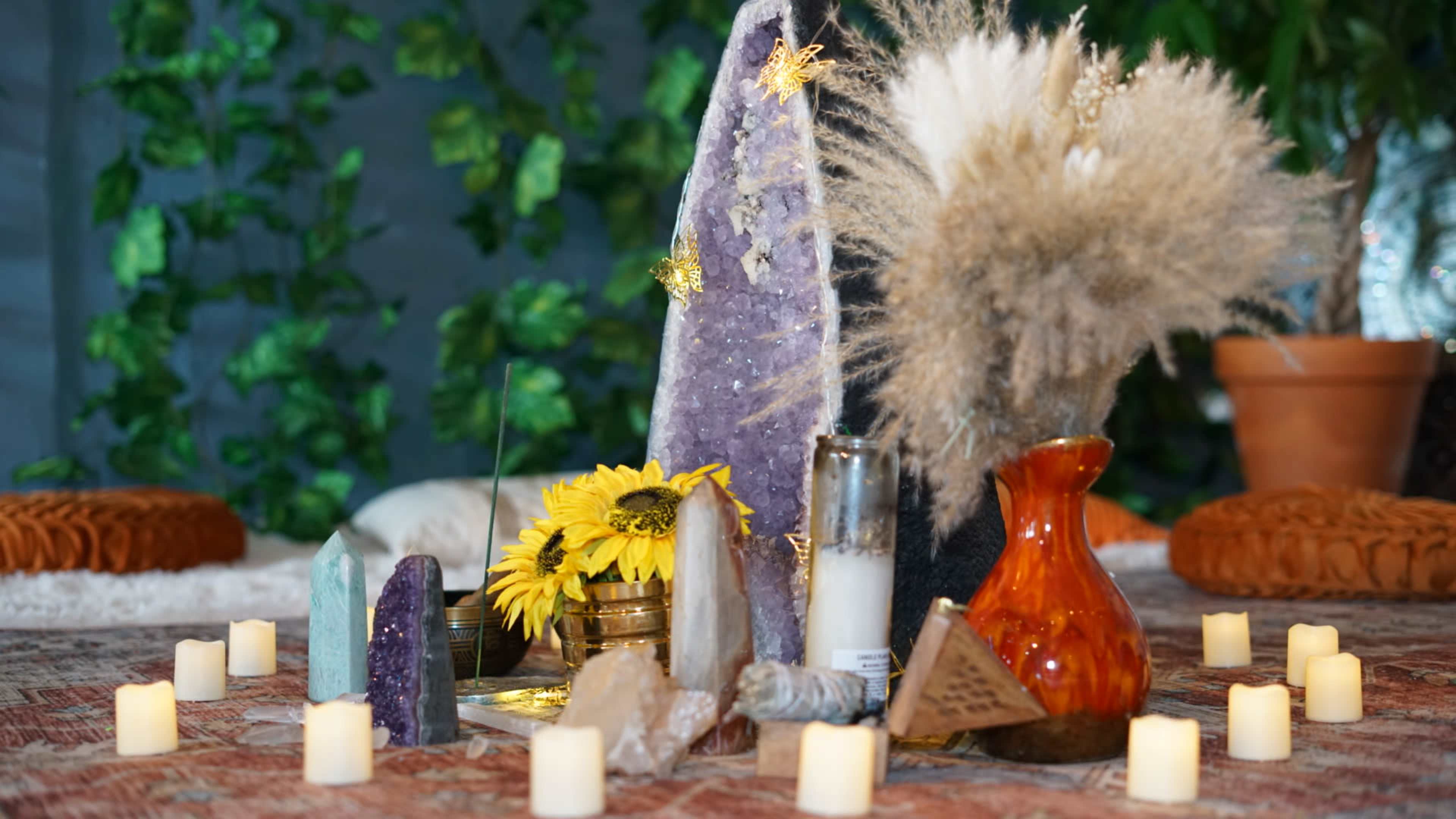A collection of crystals, dried flowers, and candles is arranged on a table surrounded by greenery.