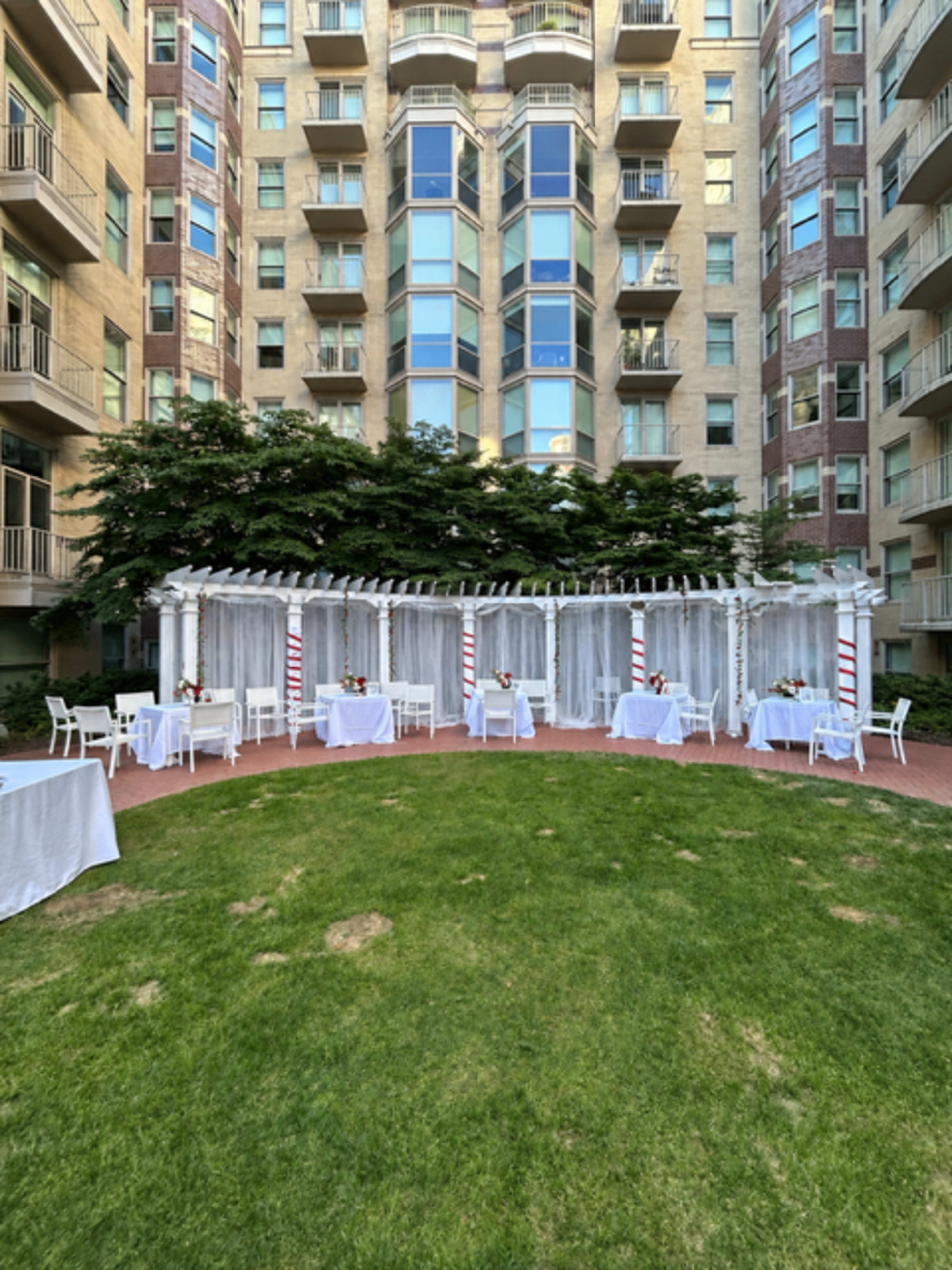 The image depicts a landscaped courtyard surrounded by a multi-story building, featuring white tables and chairs arranged under a decorative pergola with curtains.