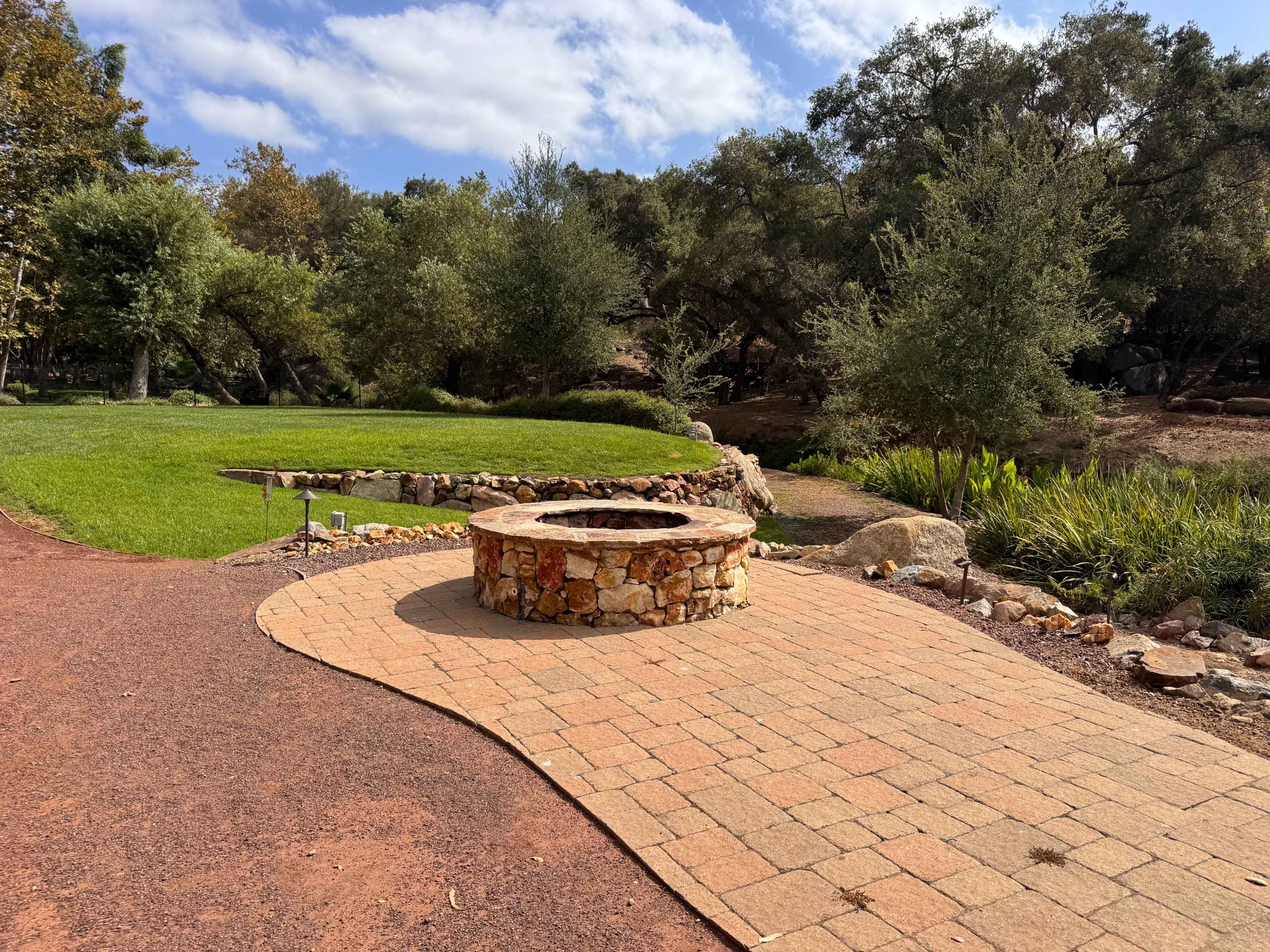 The image shows a circular stone fire pit surrounded by a paved area, with a landscaped garden featuring trees and greenery in the background.