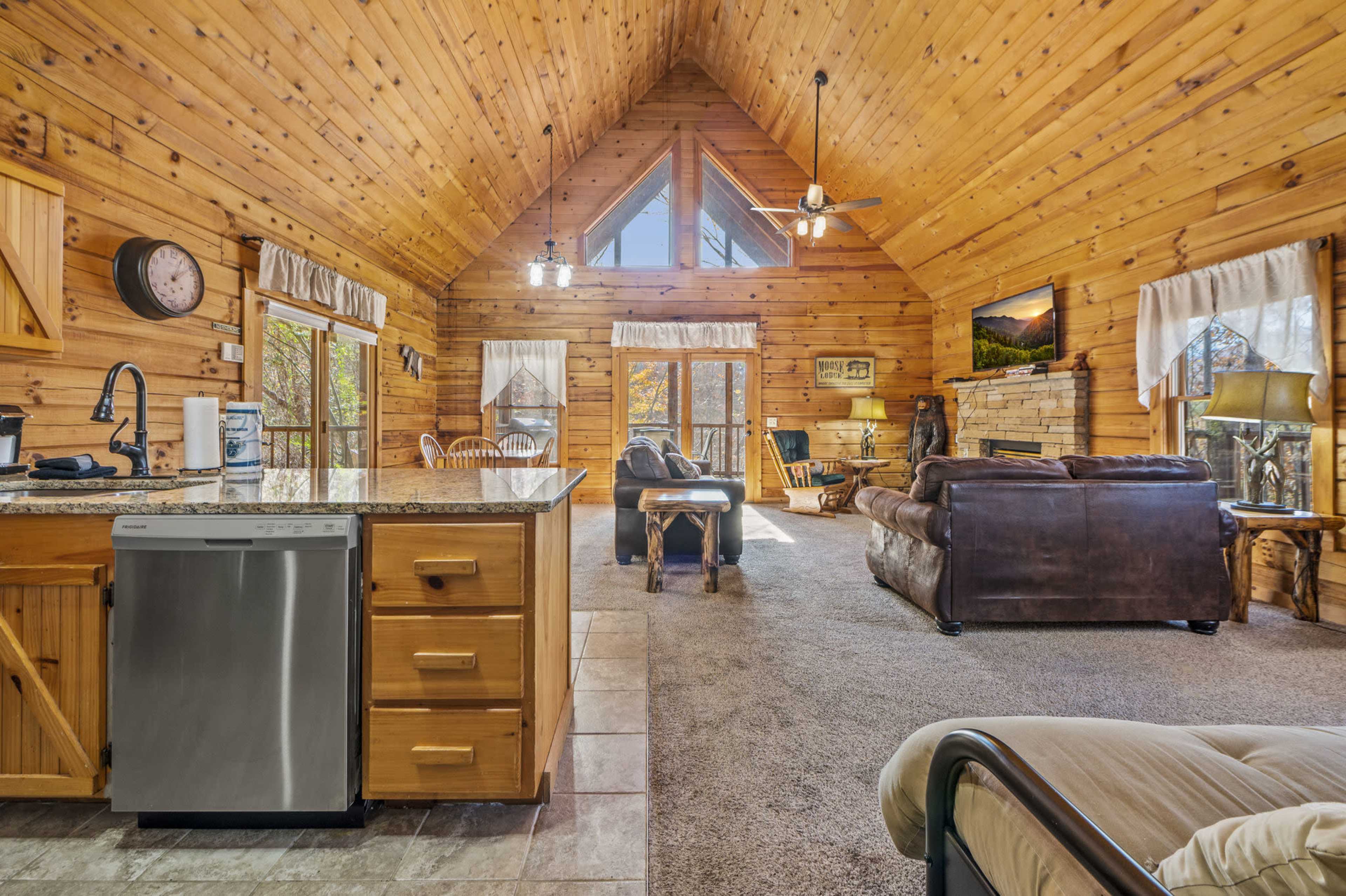 The image shows a rustic wooden interior of a cabin featuring a kitchen area, a living space with a sofa, and large windows that let in natural light.