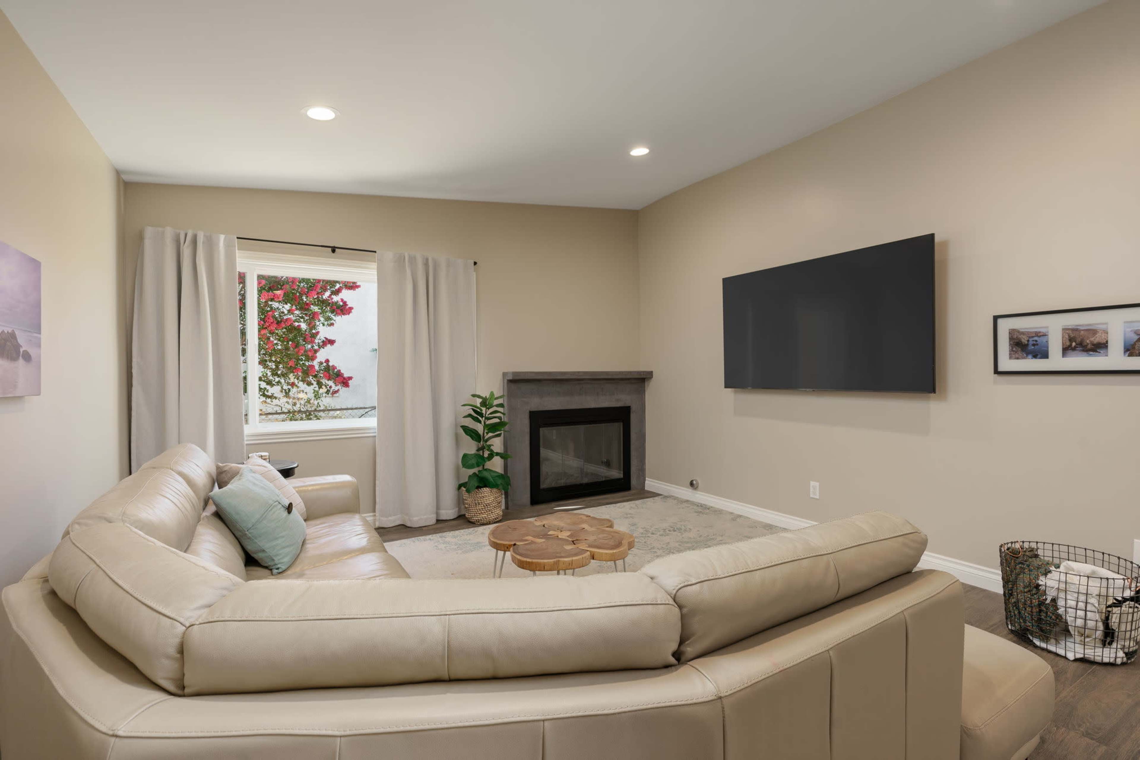 A modern living room features a beige sectional sofa, a wall-mounted television, and a fireplace beneath a window adorned with sheer curtains.