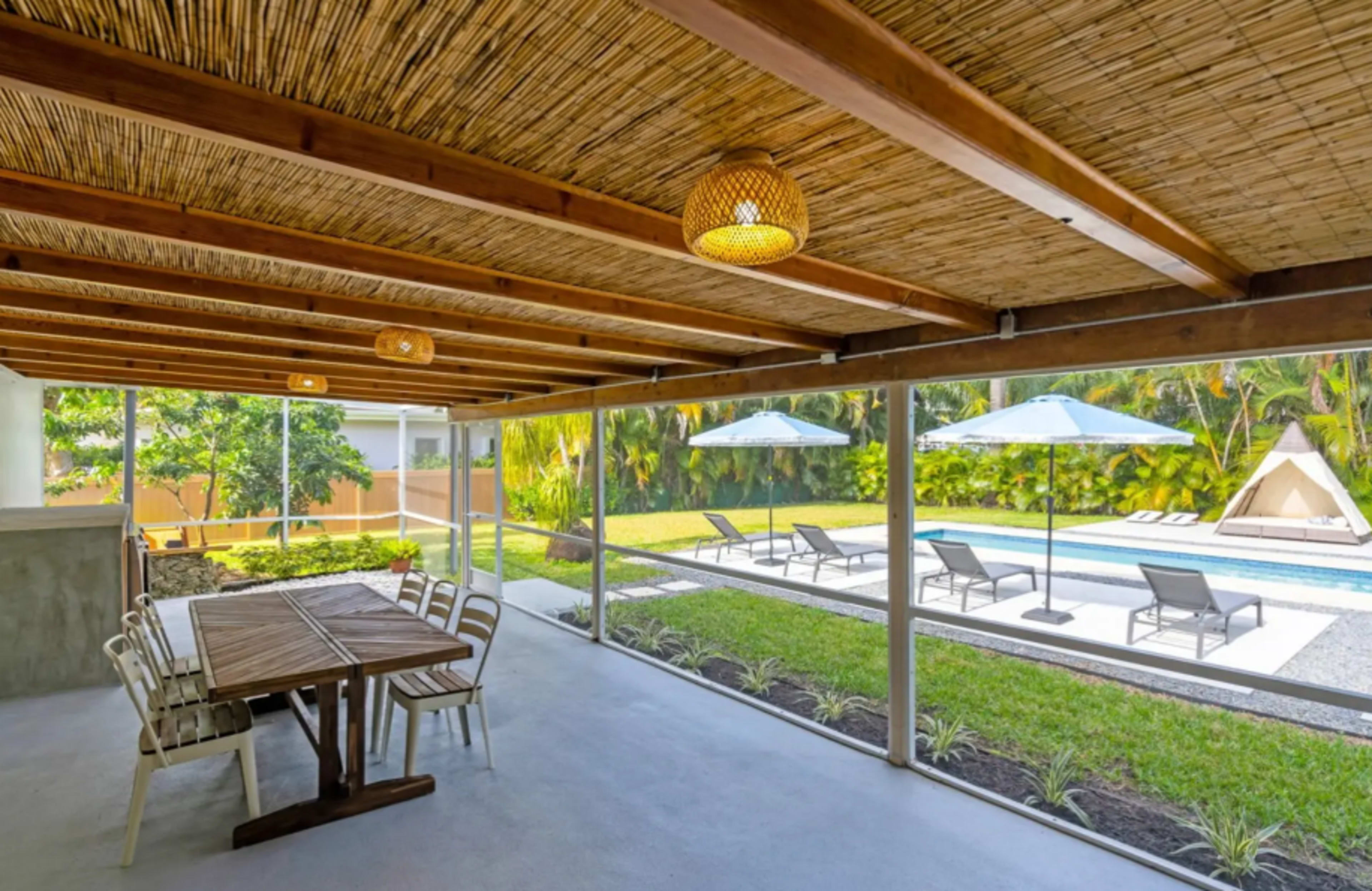 The image shows a covered patio area with a wooden dining table and chairs, overlooking a swimming pool surrounded by lounge chairs and umbrellas, set amidst a landscaped garden.