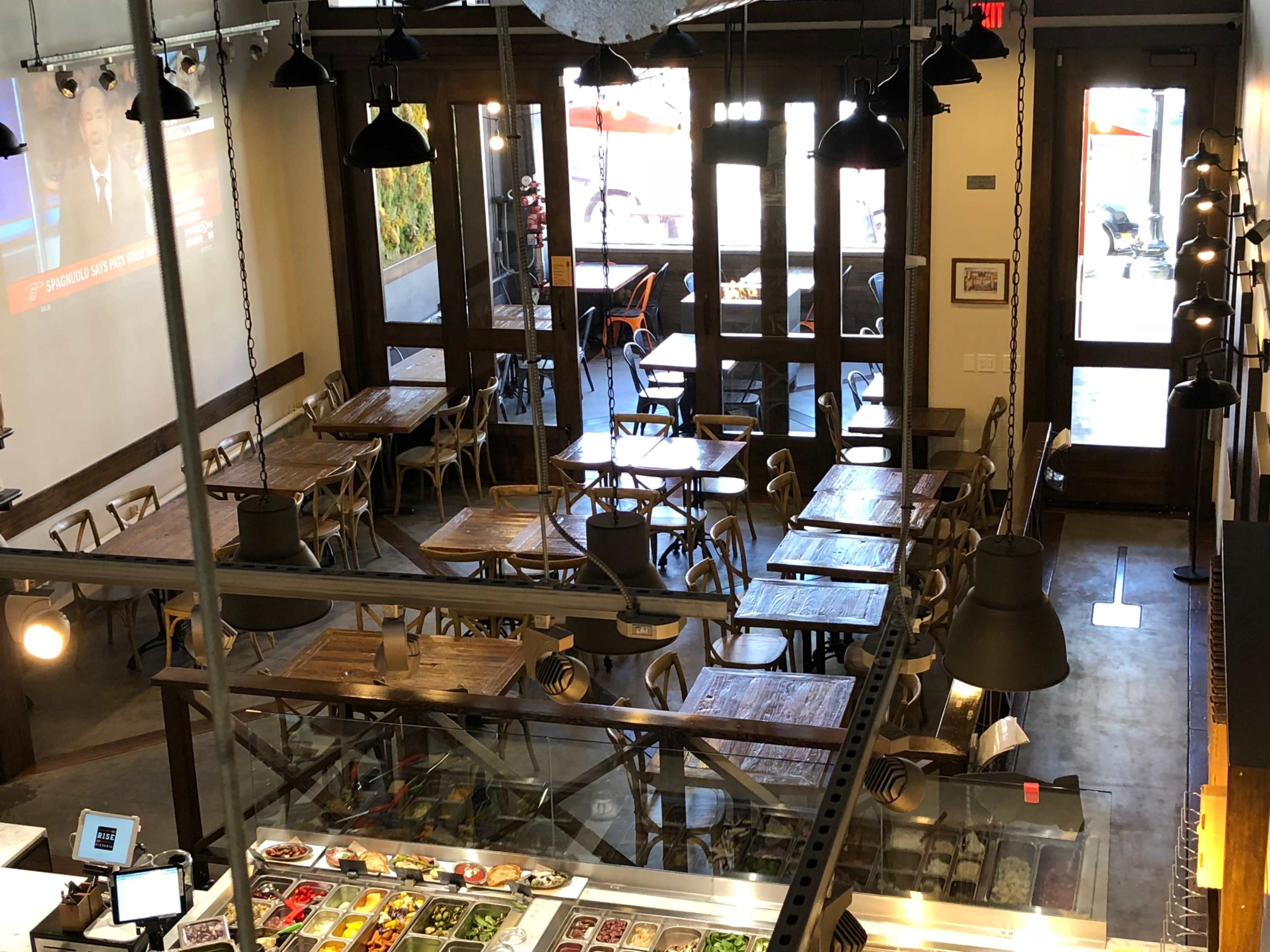 An overhead view of a cafe with wooden tables and chairs, featuring a large window and a screen displaying news.