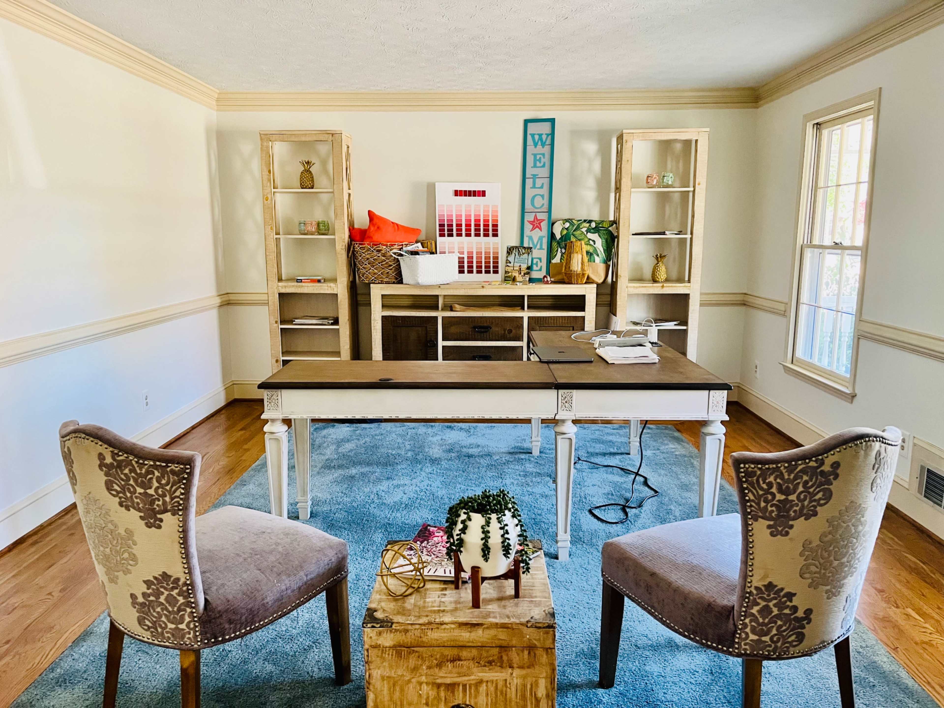 A bright home office features a wooden desk in front of two upholstered chairs, with shelves displaying decor items in the background.