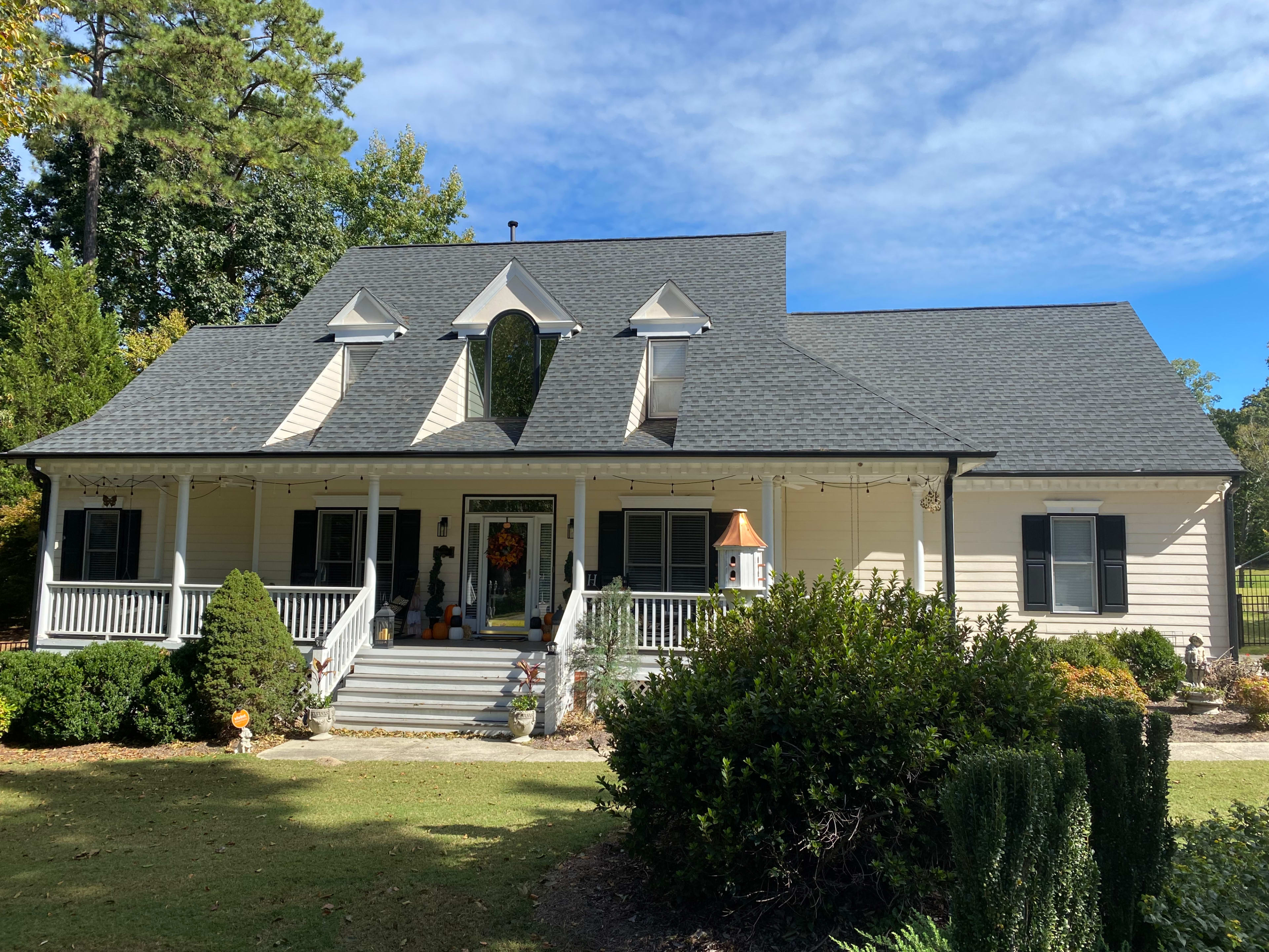 A large, light-colored house with a steep roof and a front porch is surrounded by green shrubs and trees.