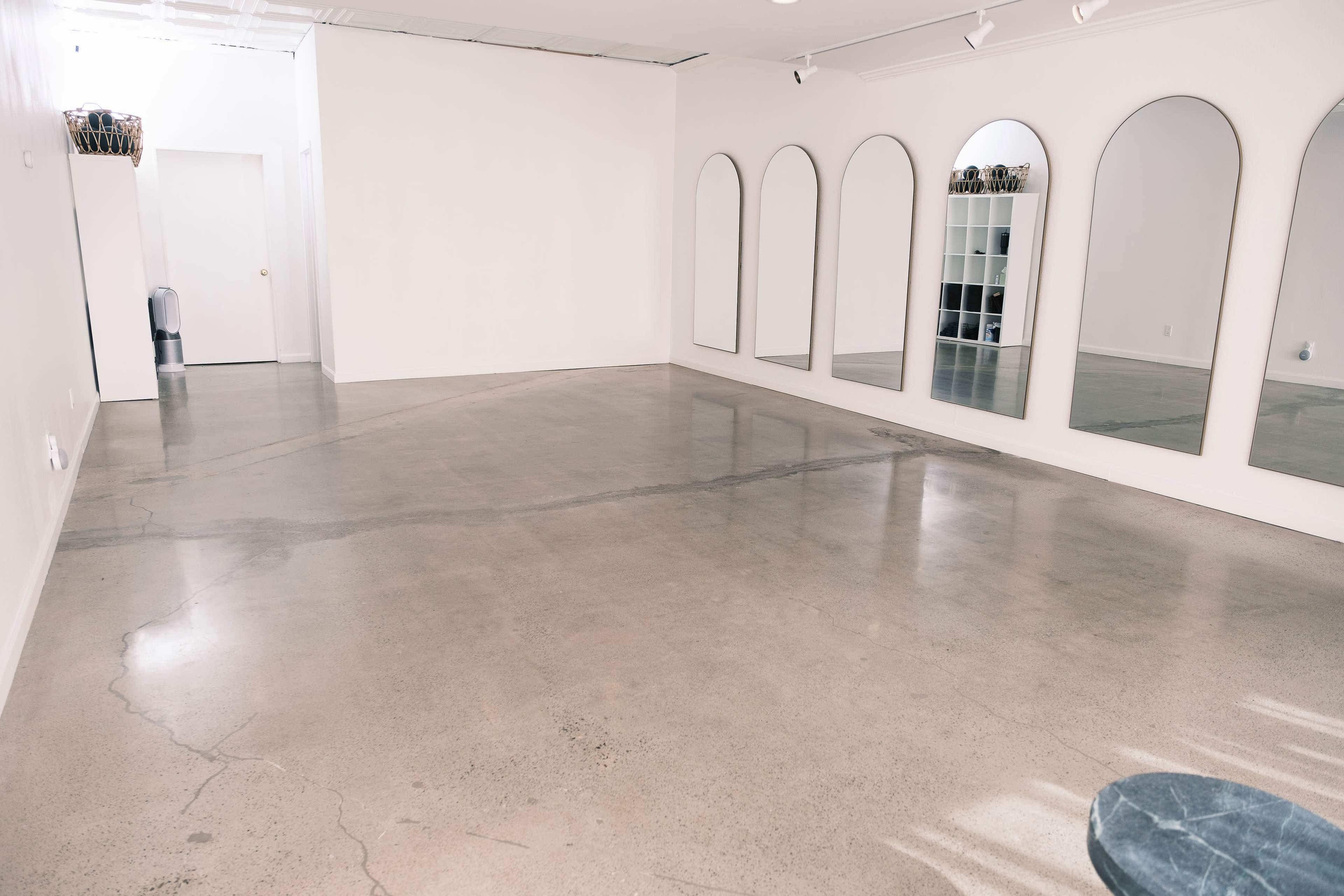 A minimalistic interior space with a polished concrete floor, large arched mirrors on one wall, and a doorway leading to another room.