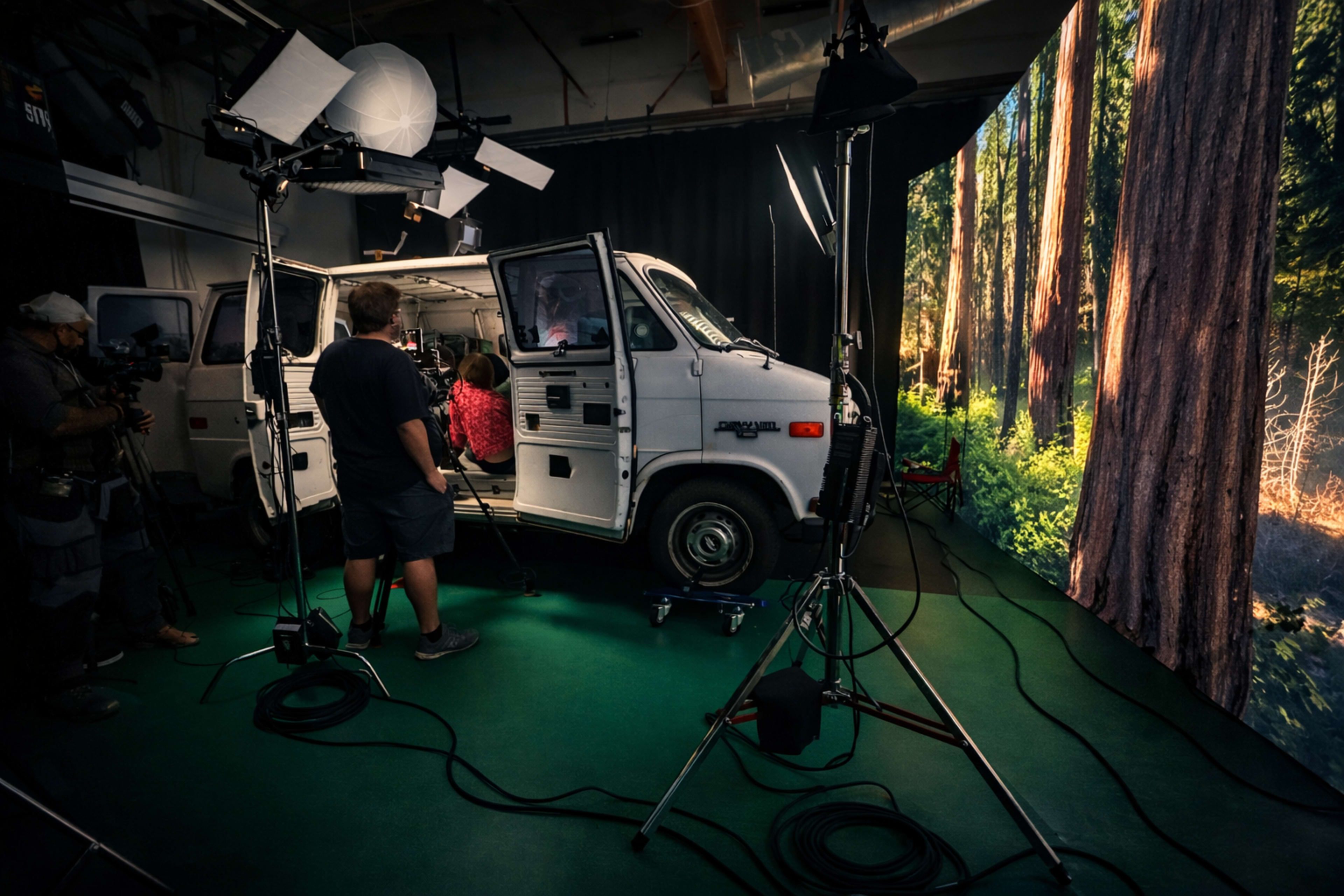 A film crew sets up equipment around a white van inside a studio, with a large forest backdrop illuminated by studio lights.