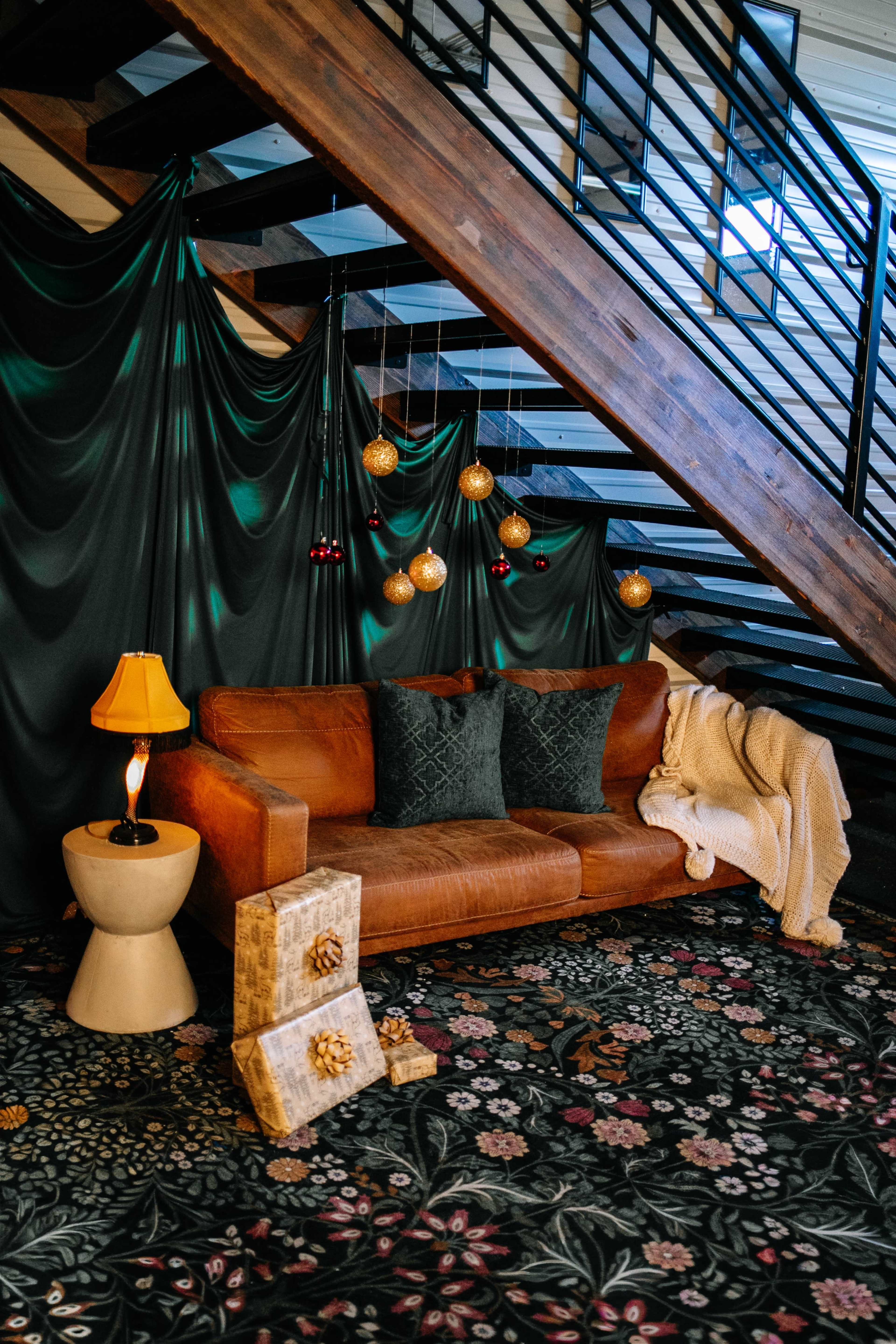 A brown leather sofa with two pillows sits on a floral carpet beside a small table lamp and decorative storage boxes, all under a staircase draped with dark fabric and adorned with hanging ornaments.