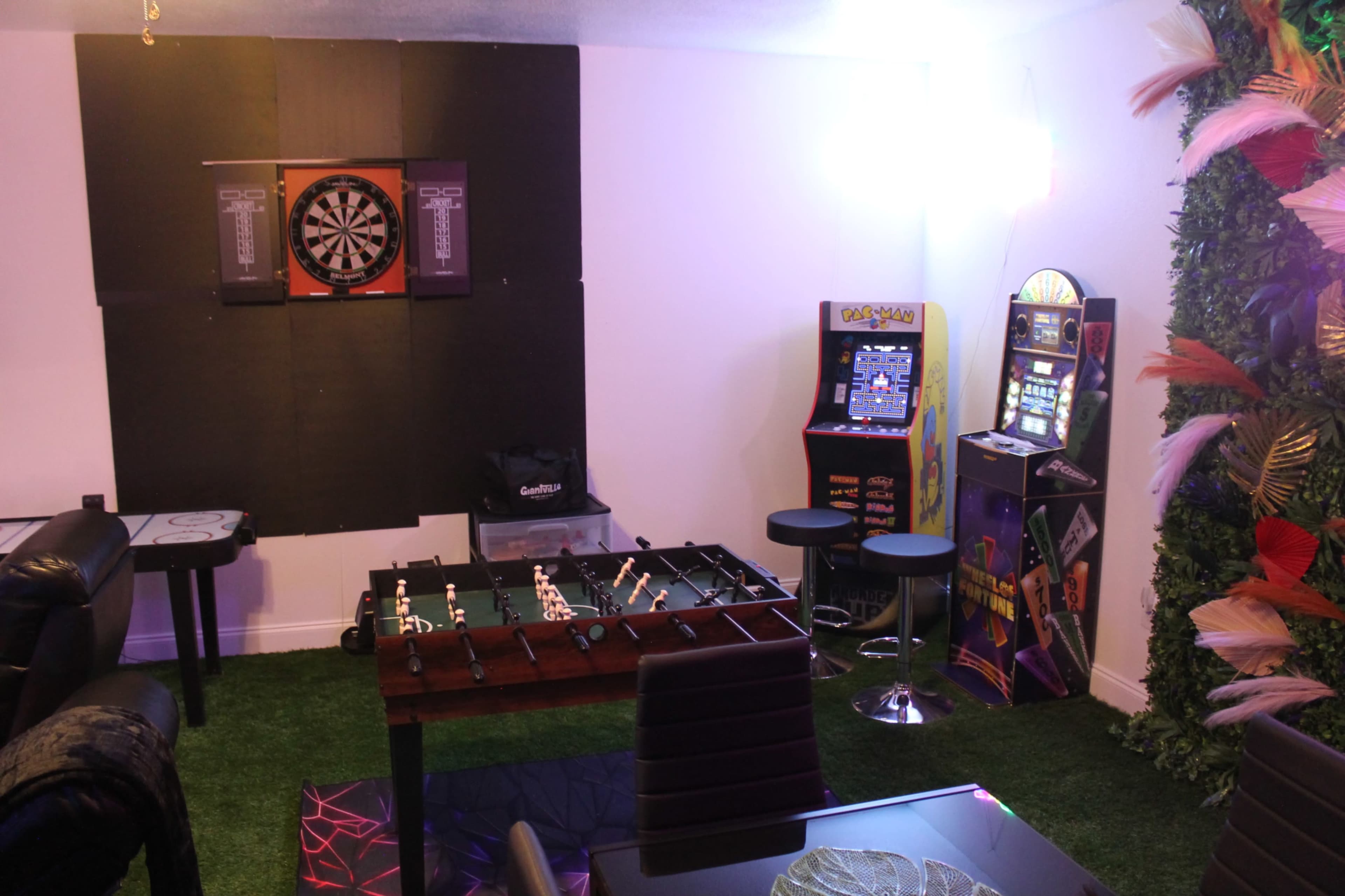 The room features a dartboard on the wall, an arcade game, a foosball table, and a synthetic grass floor.
