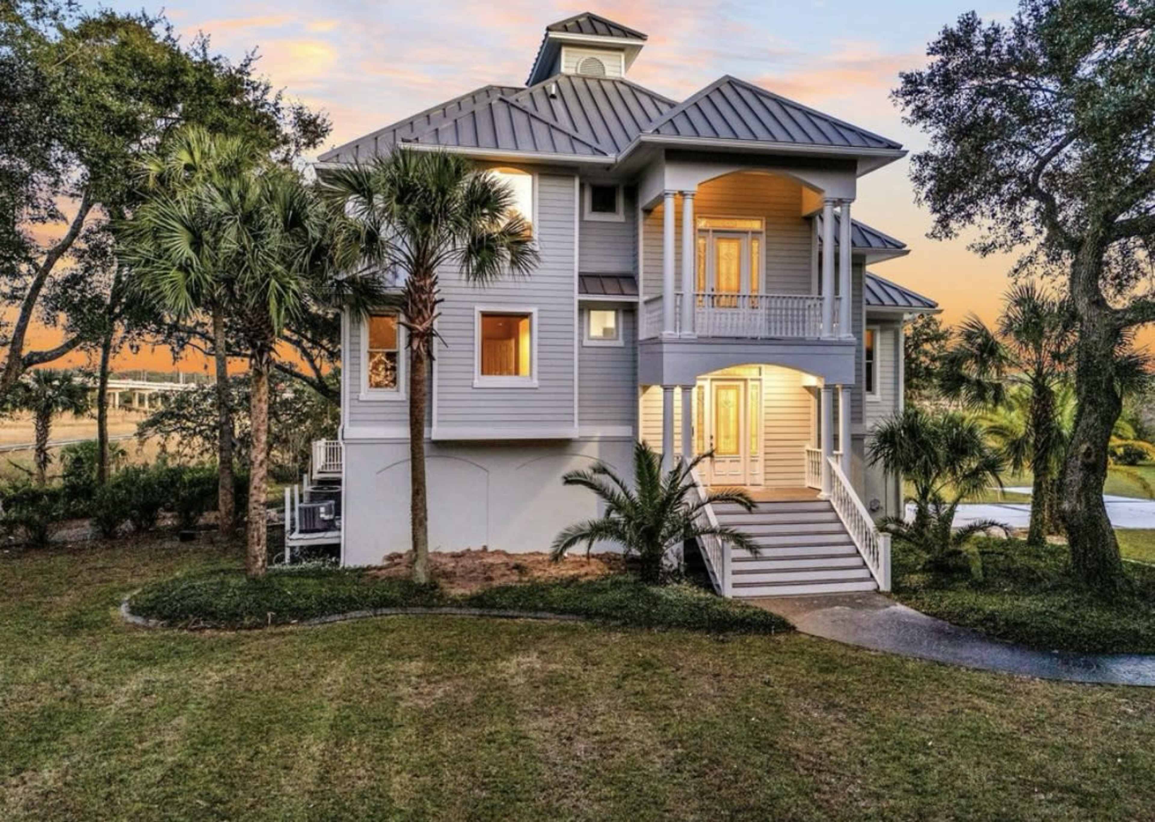 A large, two-story coastal home with a metal roof is surrounded by palm trees and green landscaping at sunset.