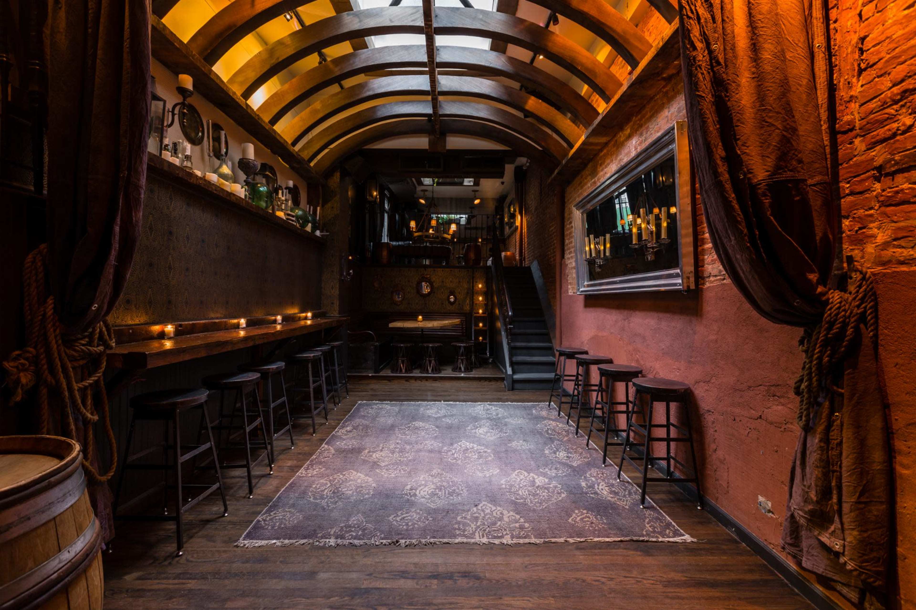 A dimly lit bar interior with a high arched ceiling, wooden bar stools along the counter, and a staircase leading to an upper level.