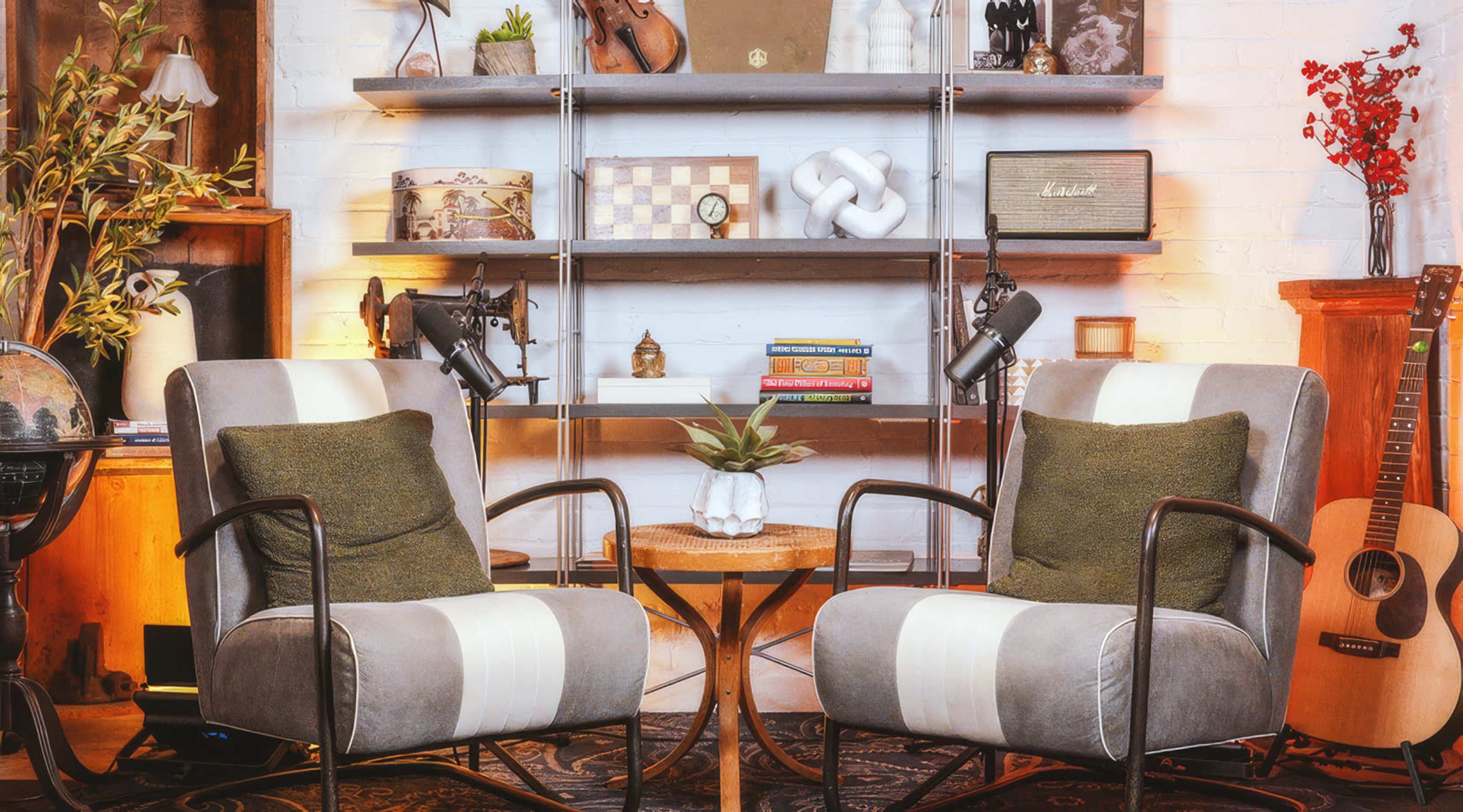 The image features two upholstered chairs with decorative pillows positioned around a small wooden table, set against a backdrop of shelves displaying various decorative items and a guitar.