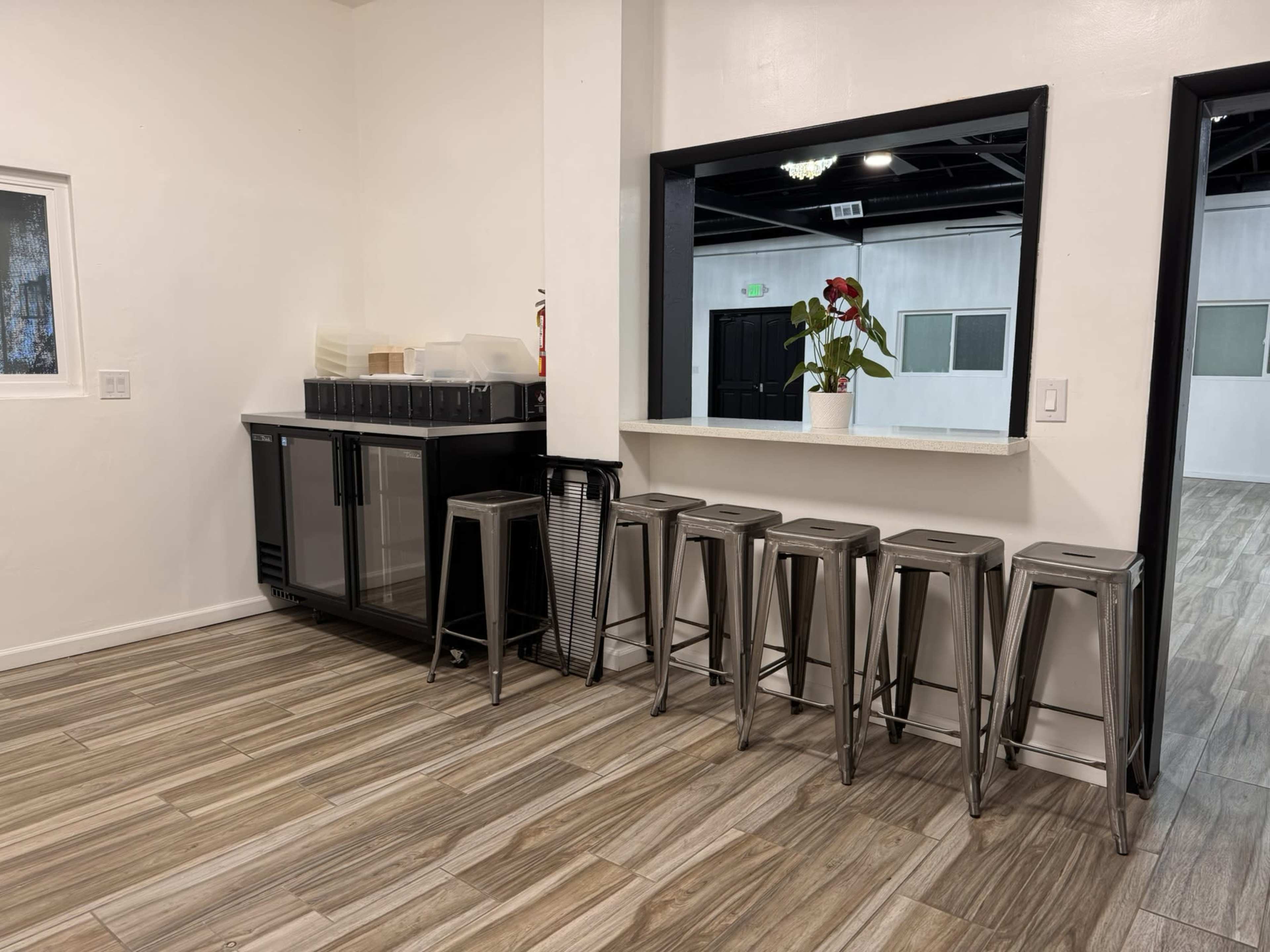 The image shows a modern interior space with a black beverage cooler, a countertop, and a row of metal stools arranged along the wall.