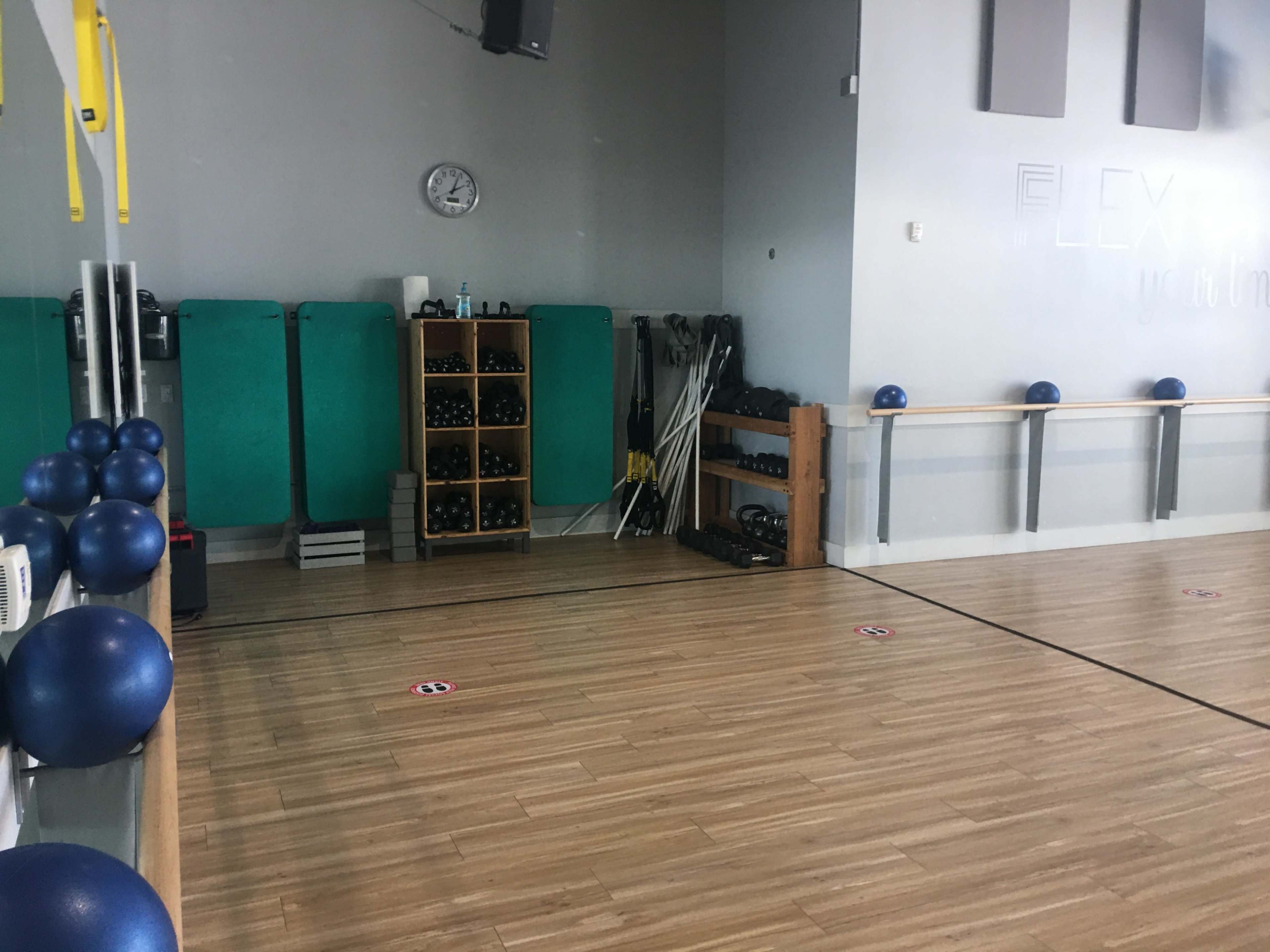 An empty fitness studio featuring wooden flooring, exercise equipment neatly organized on shelves, and several blue exercise balls lined up along the wall.