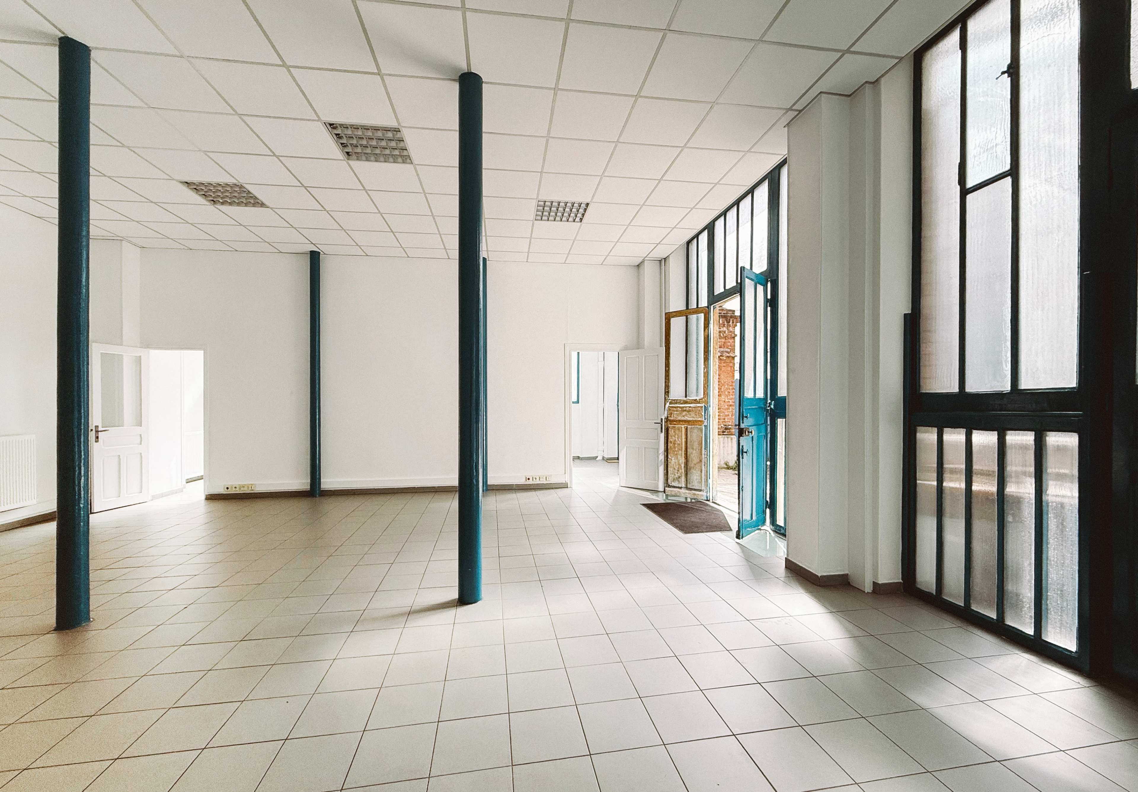 The image shows a spacious, empty room with tiled flooring, large windows, and several doorways, including one leading to an outdoor area.