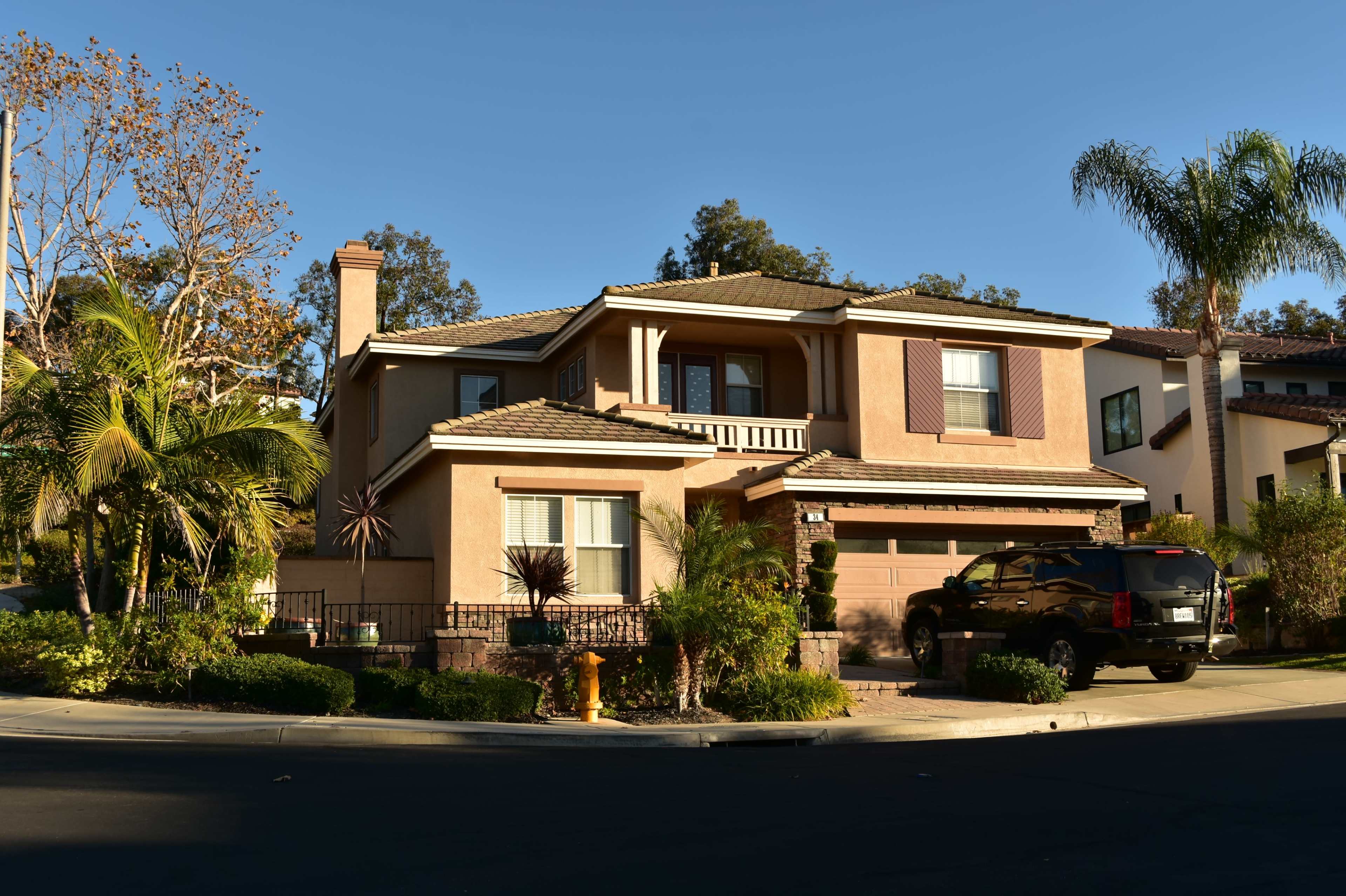 A two-story house with a tan exterior, surrounded by greenery and a palm tree, located on a corner lot in a suburban neighborhood.