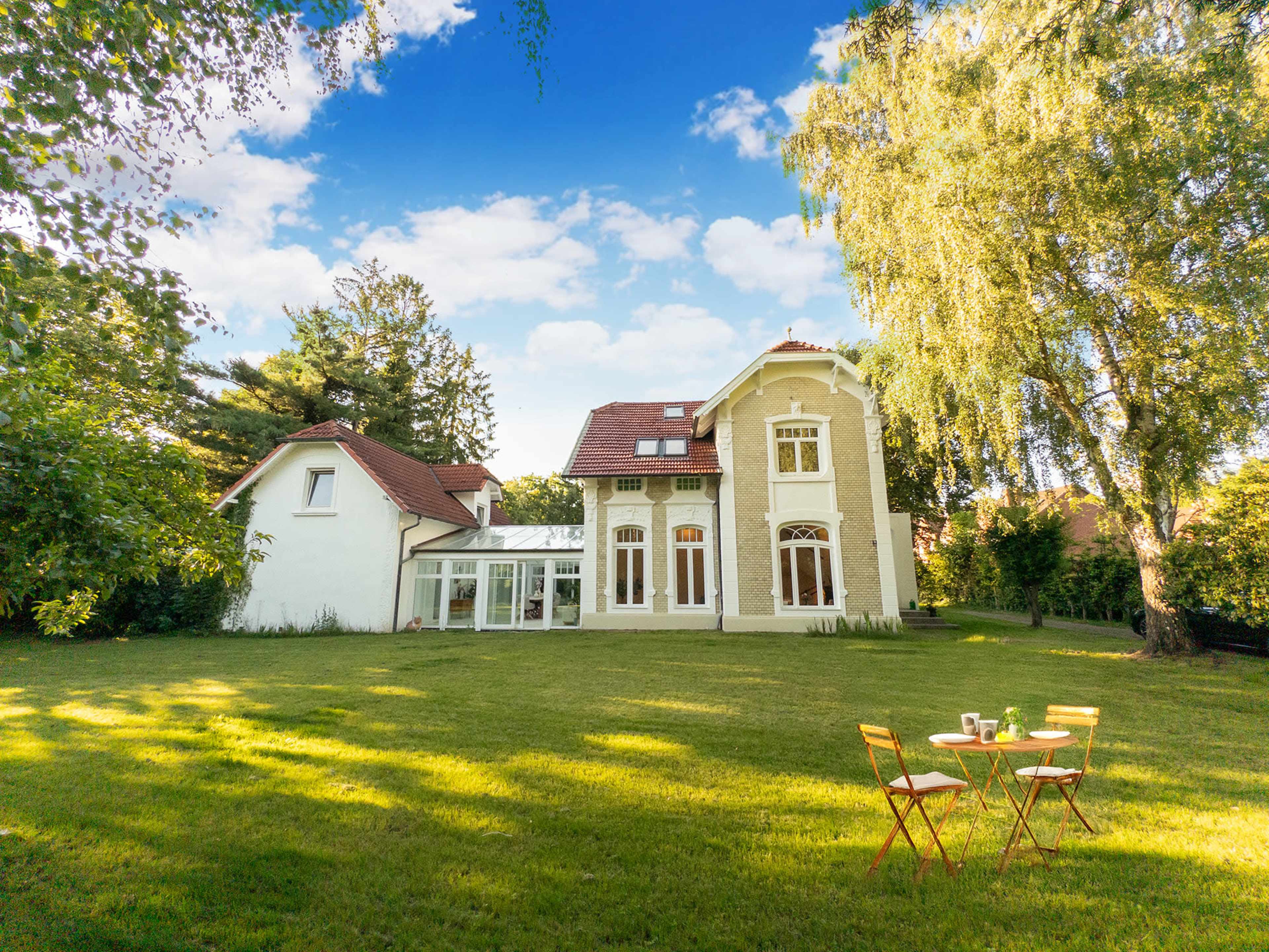 A large house with a white exterior and multiple gables stands in a spacious green backyard adorned with a small round table and chairs.