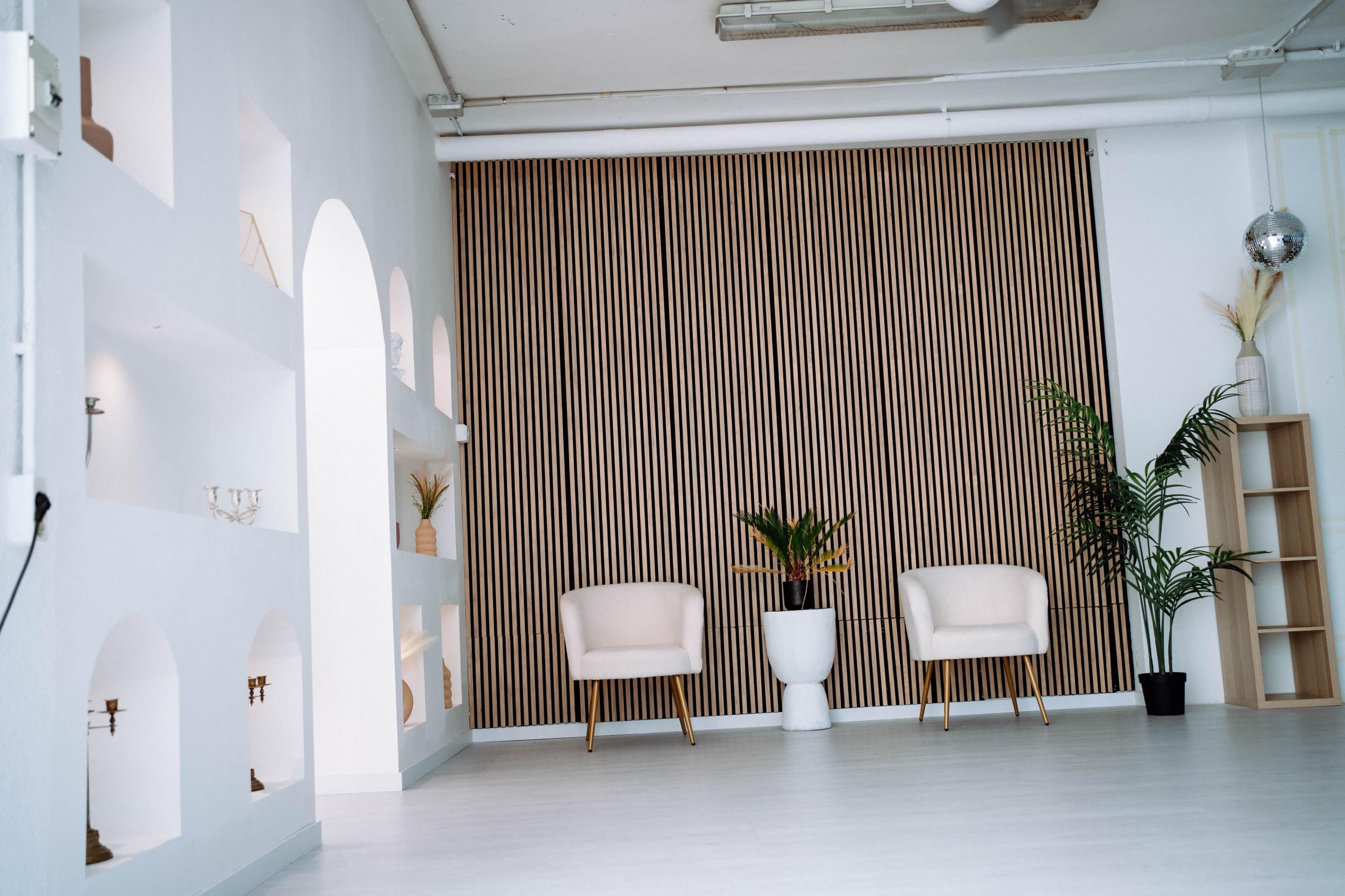 The image shows a minimalist interior with two cream-colored chairs, a plant, and a wooden slatted backdrop.