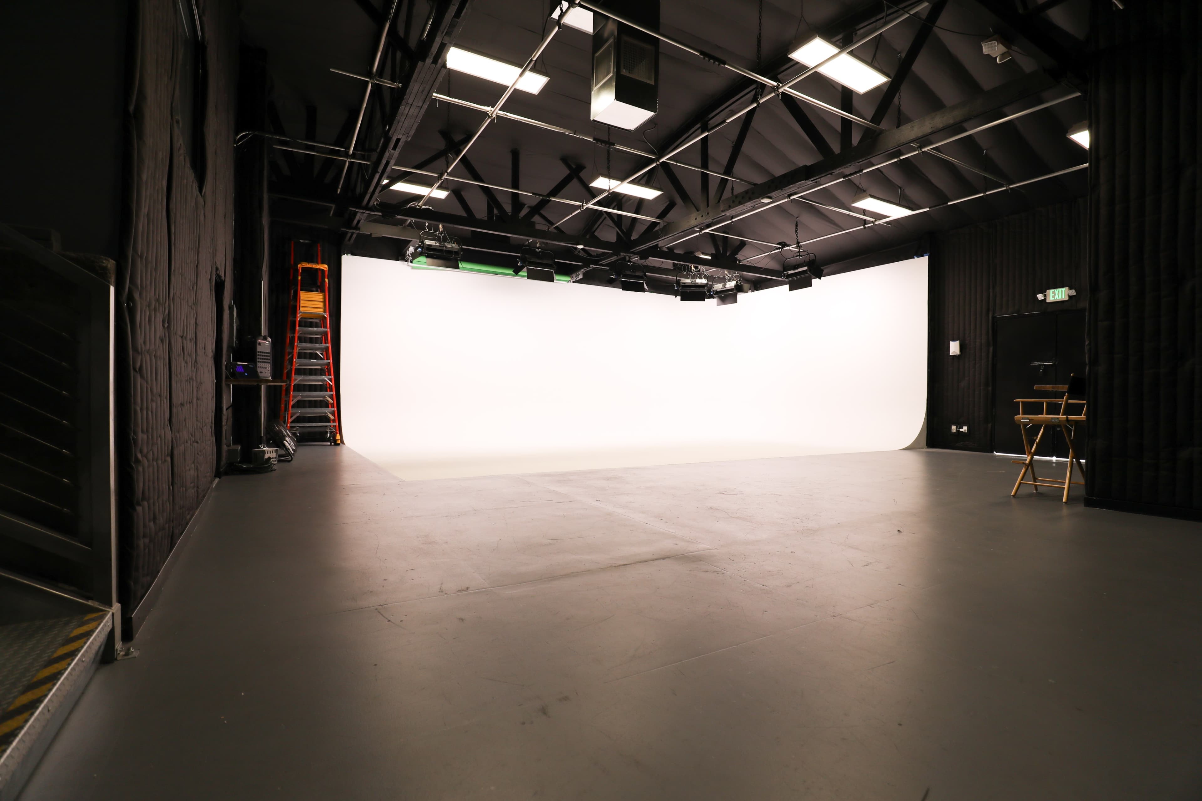 The image shows an empty studio space with a large white backdrop, overhead lighting, and ladders against the wall.