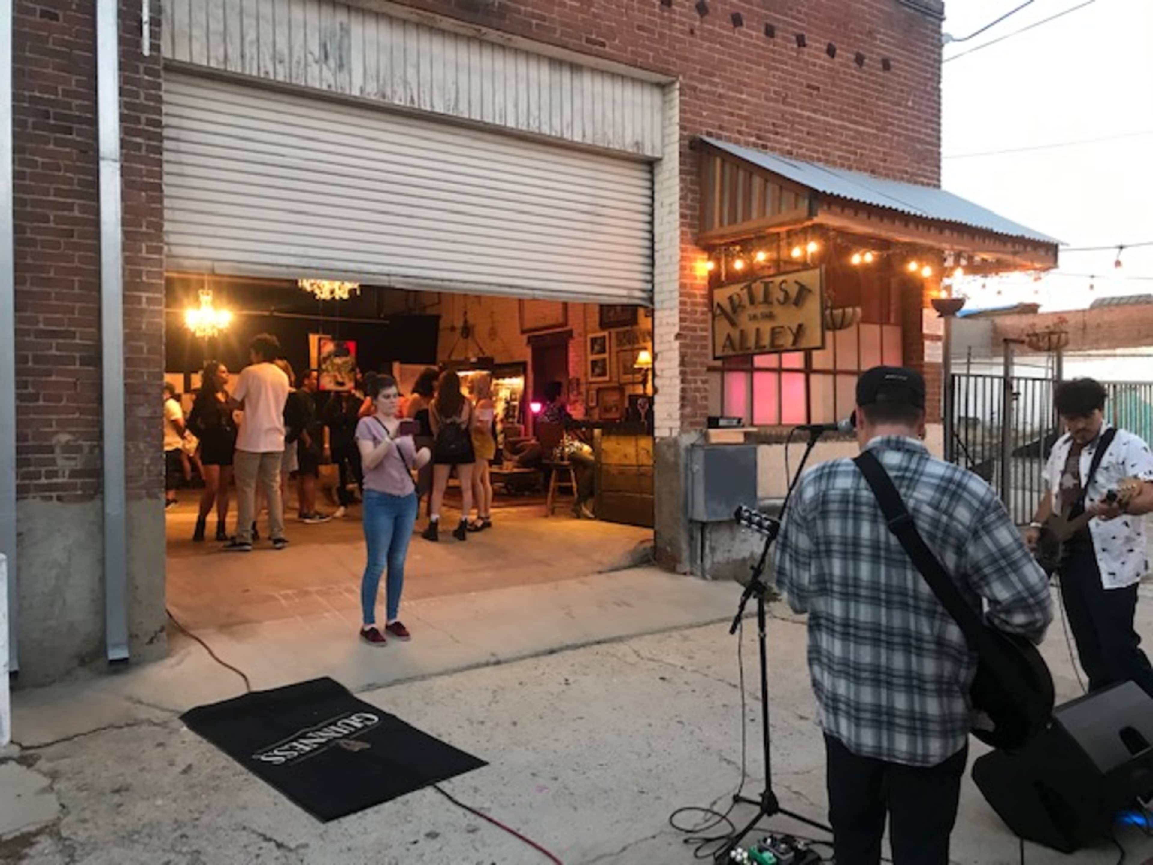 A group of people is gathered in an open alleyway for an outdoor music event, with a band playing inside a warehouse-like space adorned with colorful lights.