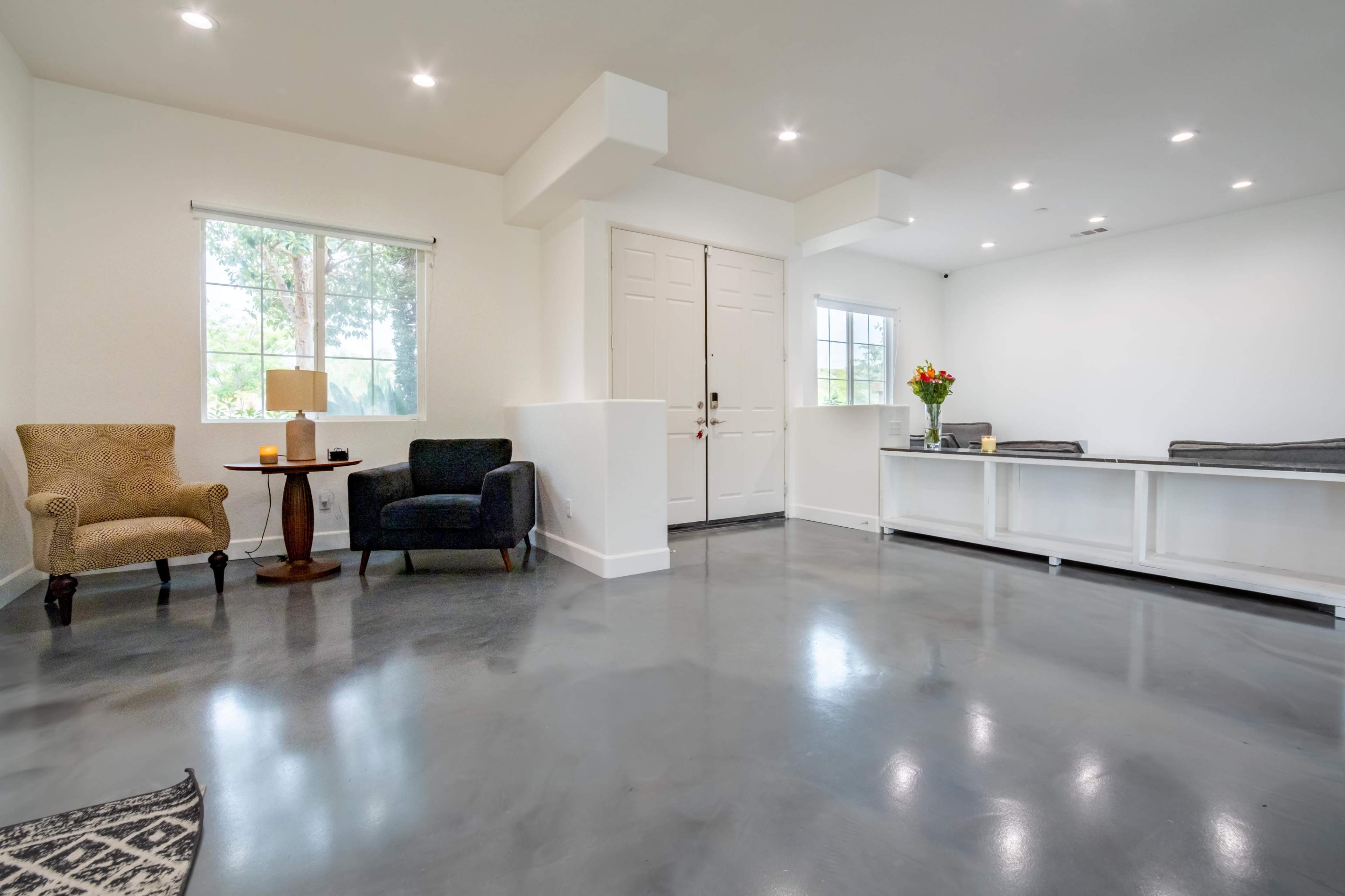 A spacious room features two chairs, a small table, and a flower arrangement by the door, with large windows providing natural light.