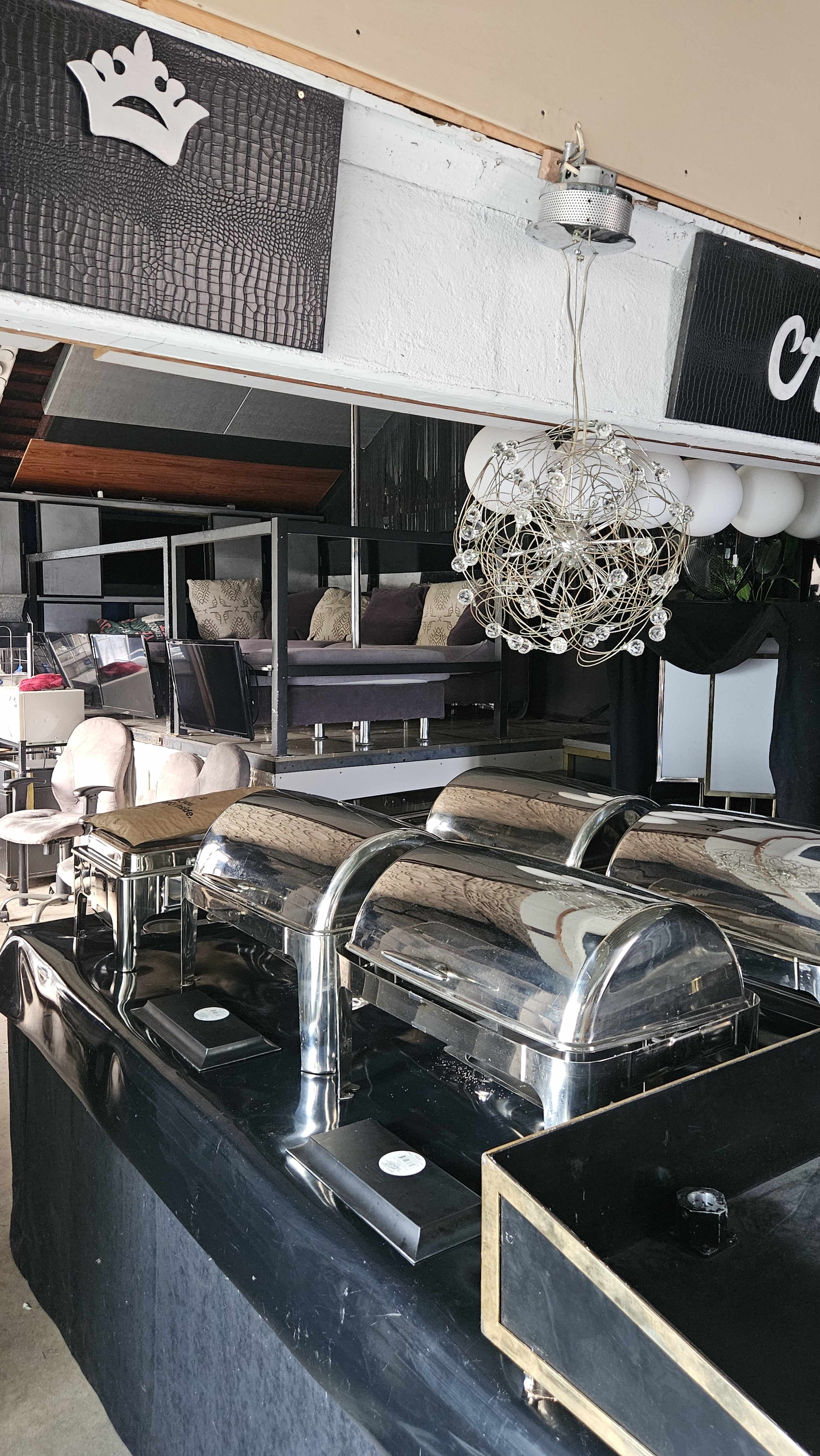 The image shows a caterer setup with metallic chafing dishes on a black table, a stylized chandelier above, and seating in the background.