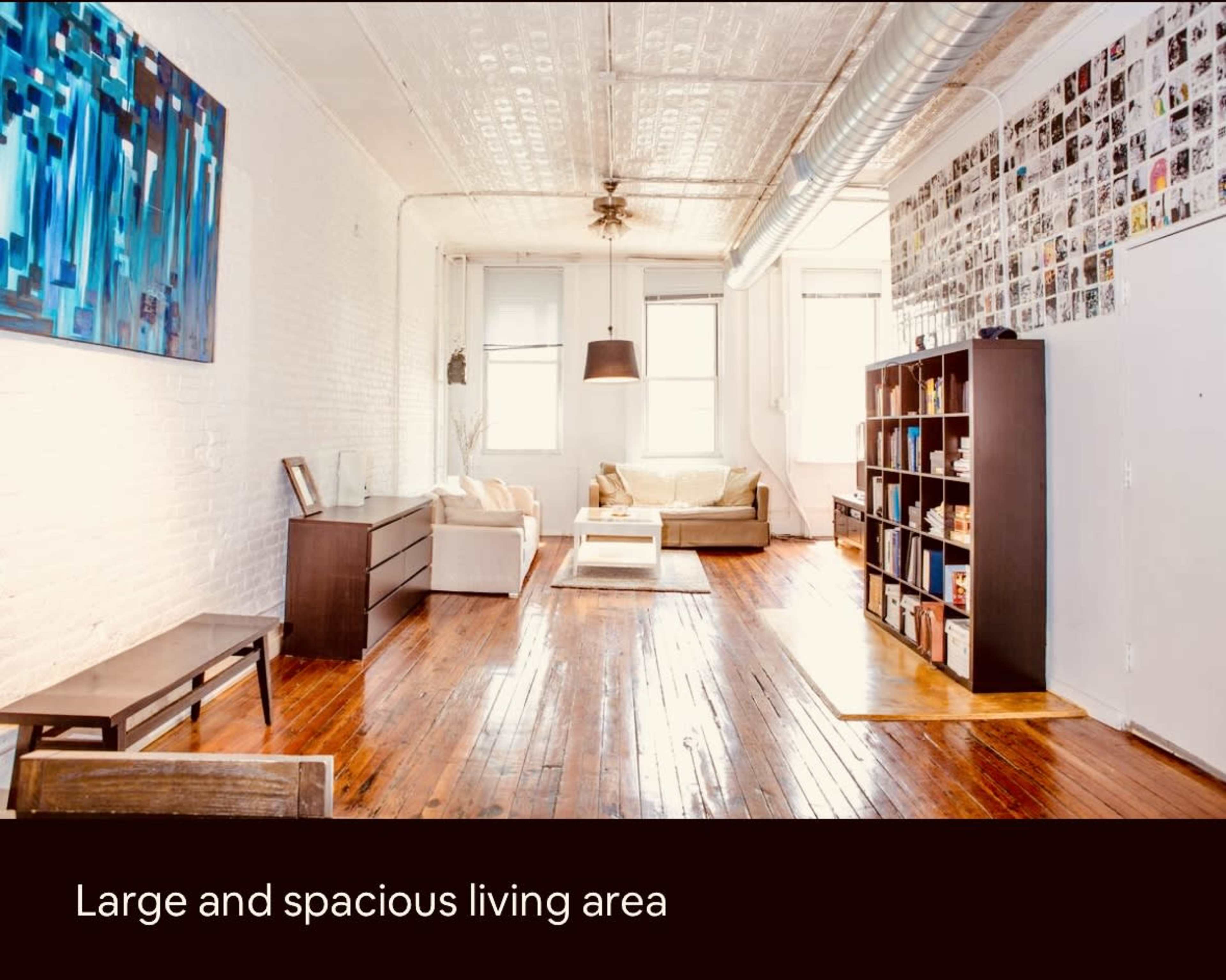 The image shows a bright living area with a wooden floor, white walls, two sofas, a bookshelf, and large windows allowing natural light to enter.