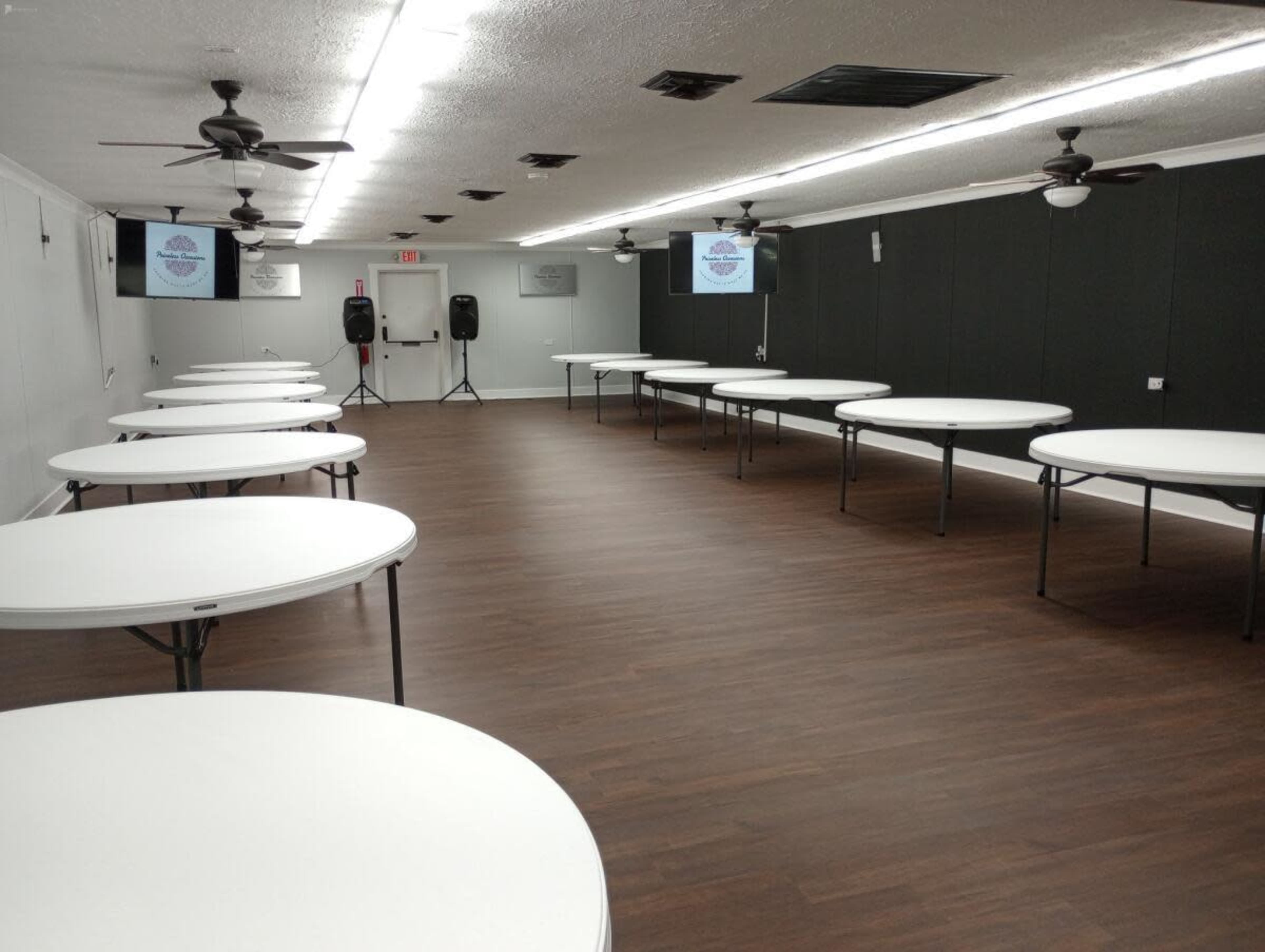 A spacious room set up with several round tables arranged for an event, featuring a wooden floor and ceiling fans.