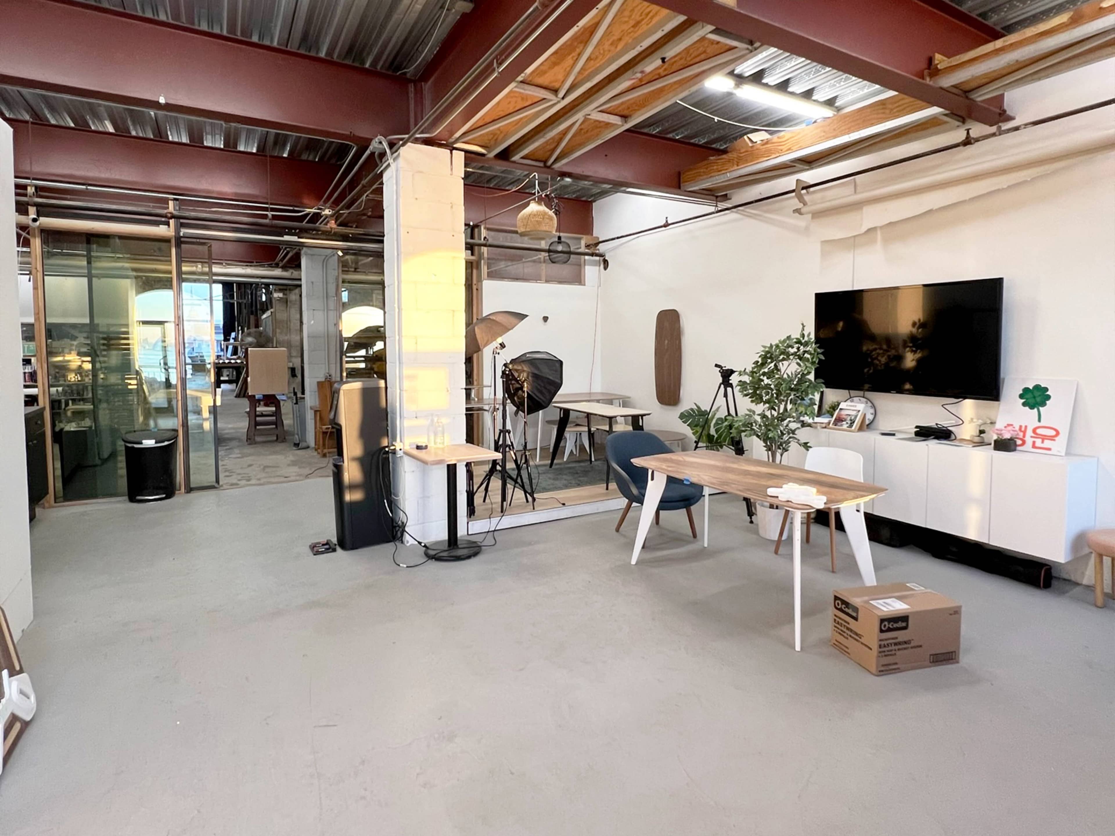 The image features a spacious, minimalist office with a wooden table, chairs, and various equipment set up for photography or videography.