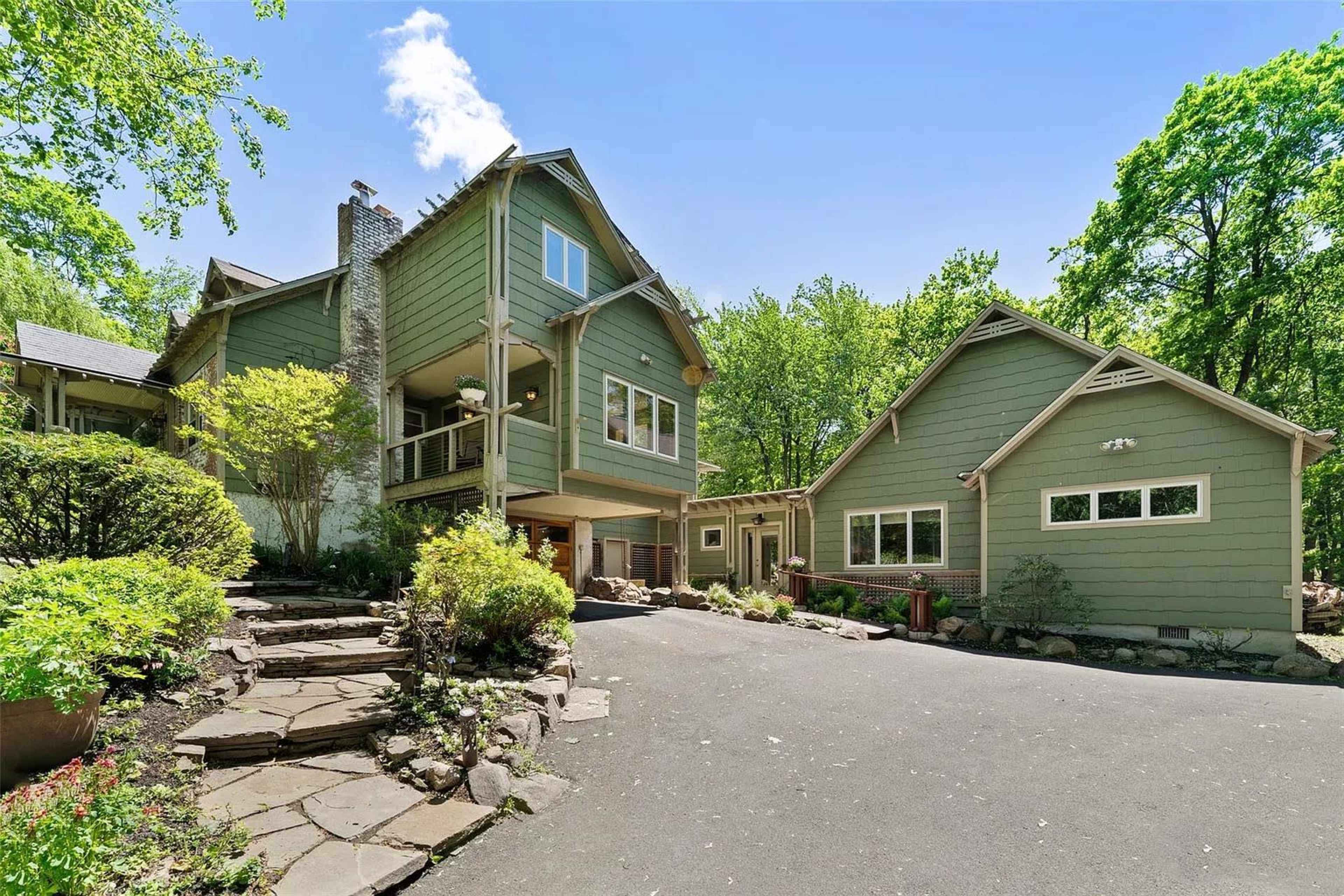 A large green house with multiple levels is surrounded by trees and features a stone pathway and a paved driveway.