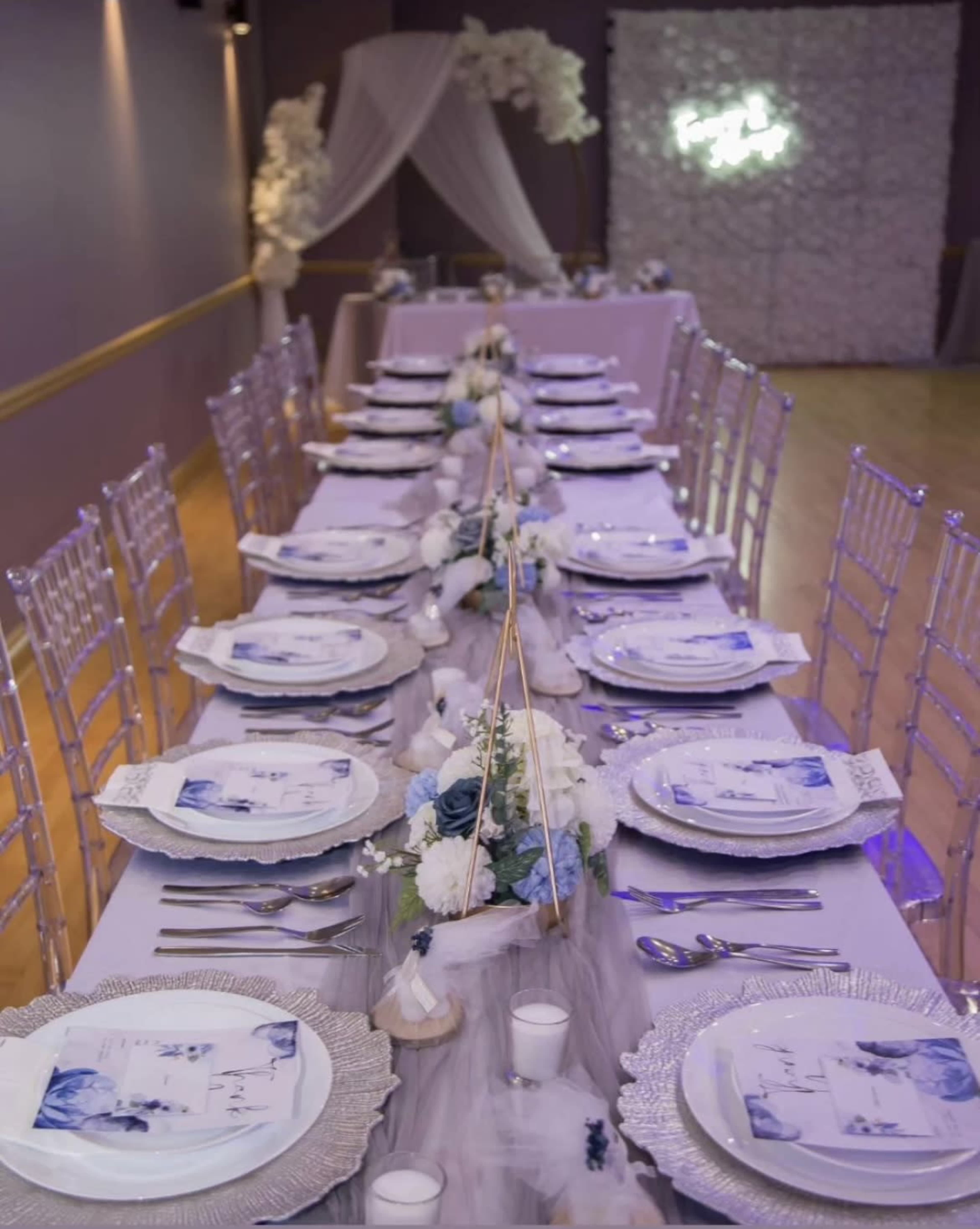 A long banquet table is elegantly set with white tableware and floral arrangements, illuminated by soft lighting and decorative accents.